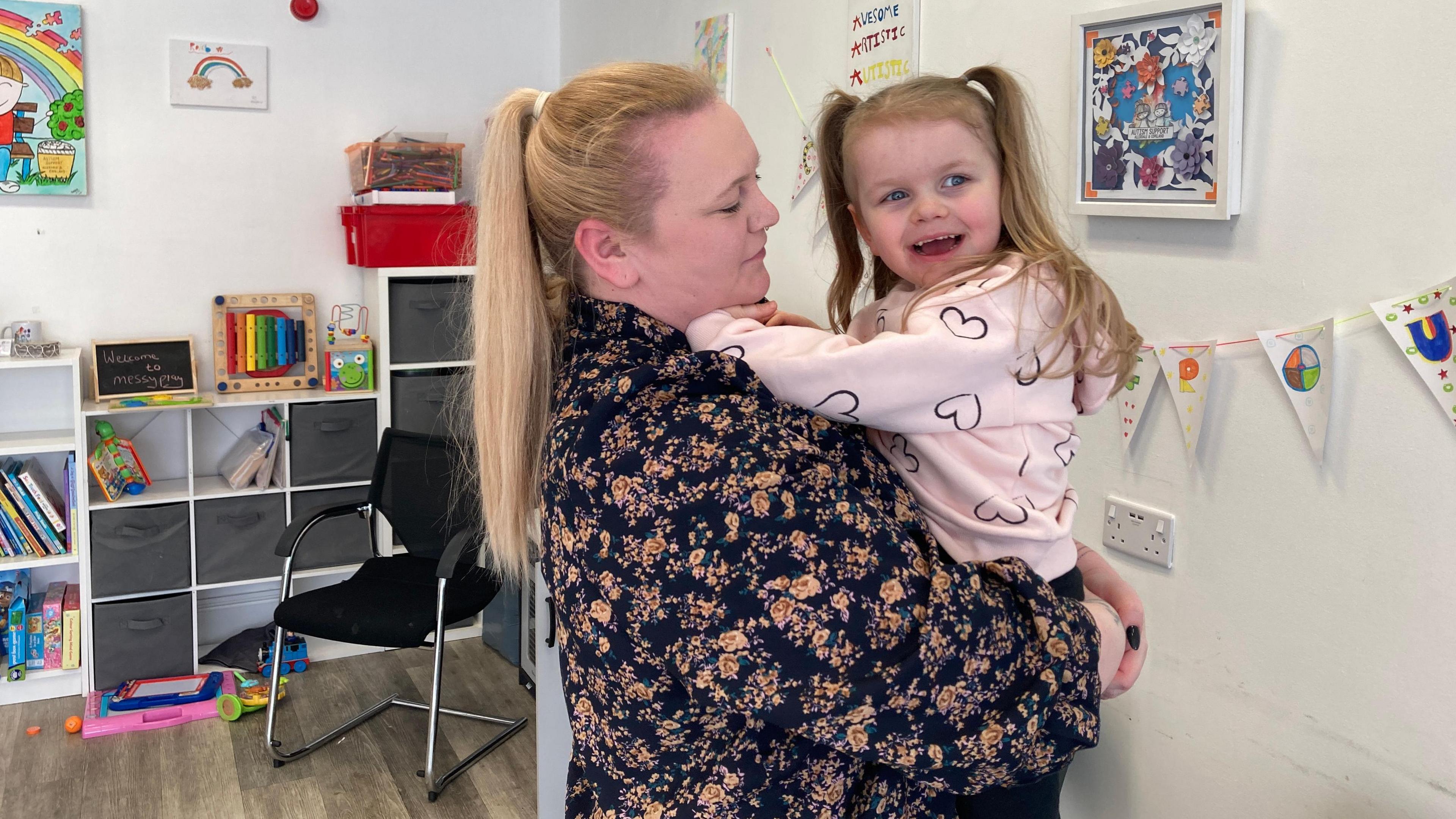 Melanie Askew is holding her daughter in her arms in a room with lots of toys and flags on the wall. Melanie has a flowery top on and her long blonde hair is tied back. The child has her hair in bunches and is laughing.