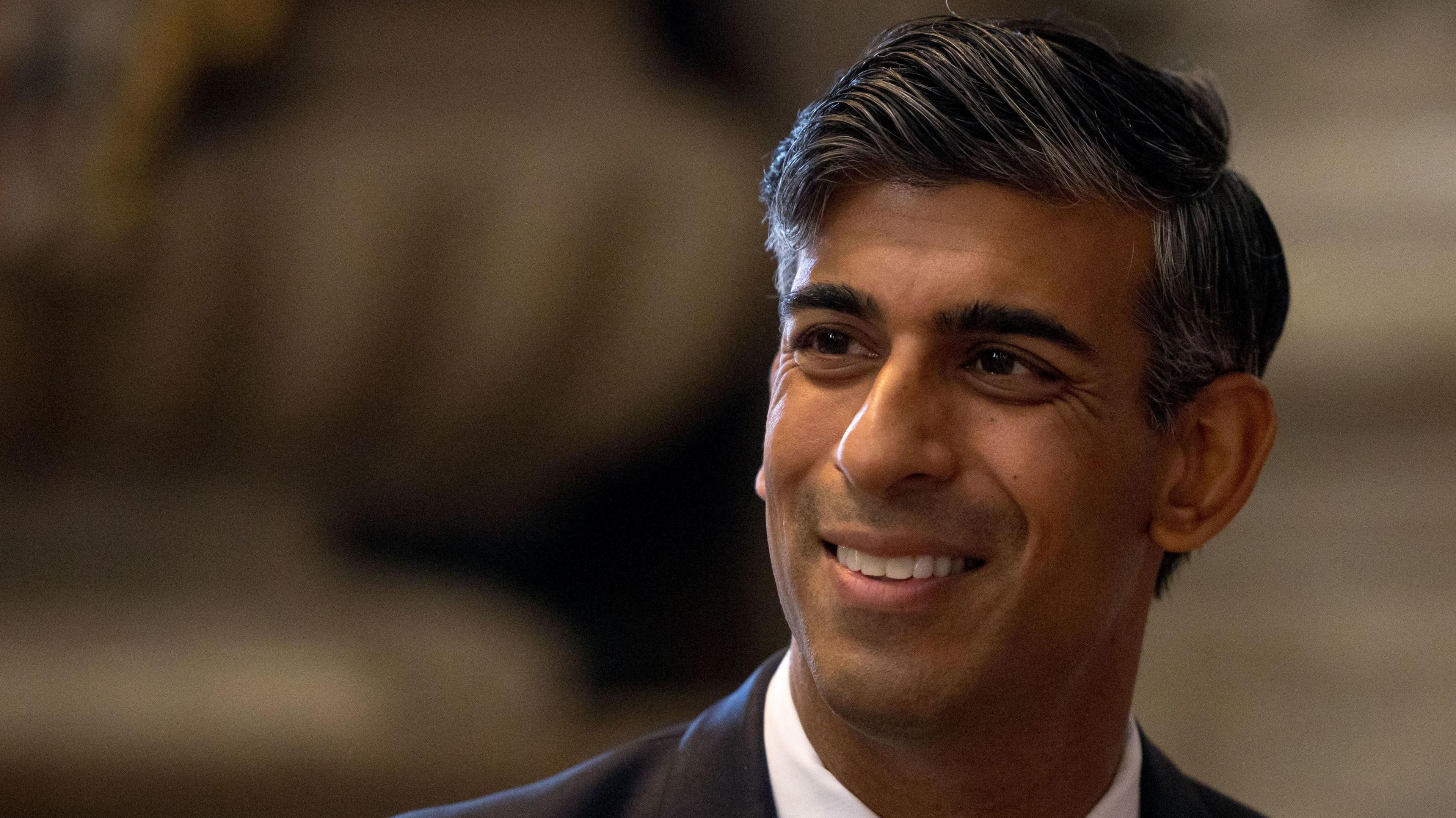 Rishi Sunak - he has black hair, is smiling and is wearing a suit. 