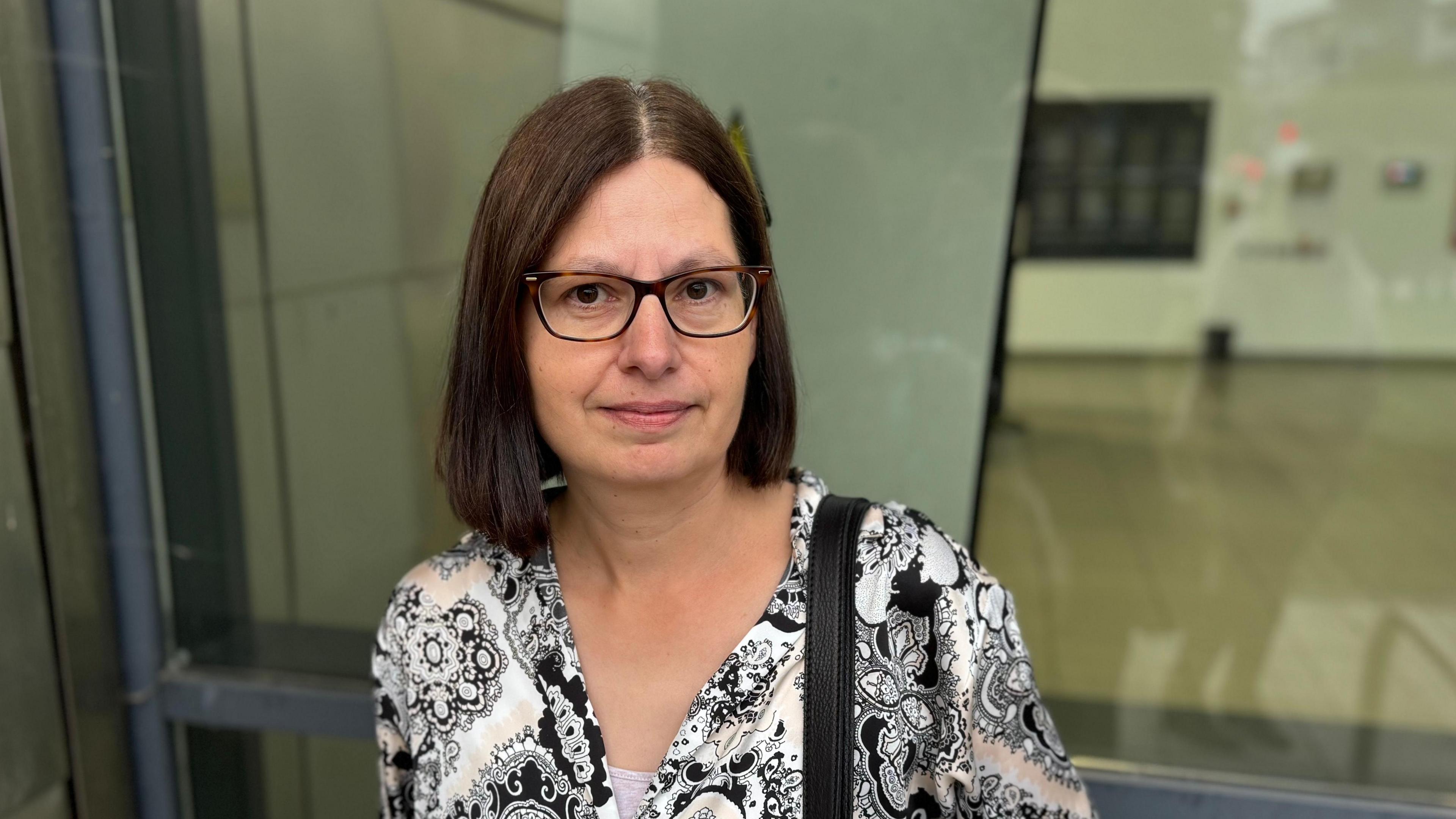 A woman with dark brown shoulder length hair wearing glasses. She has a black and white patterned top on and is standing in front of a window.