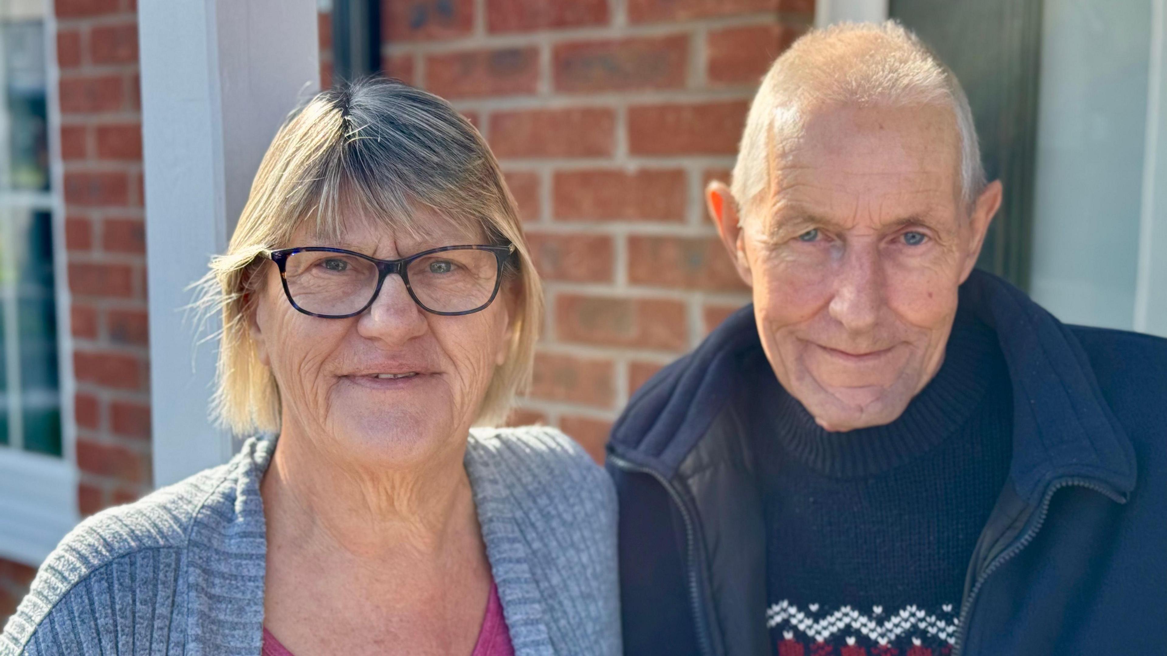 A woman with short blonde hair and black glasses is standing next to a man with short grey hair and a blue jumper and jacket. They are in front of a brown brick house.