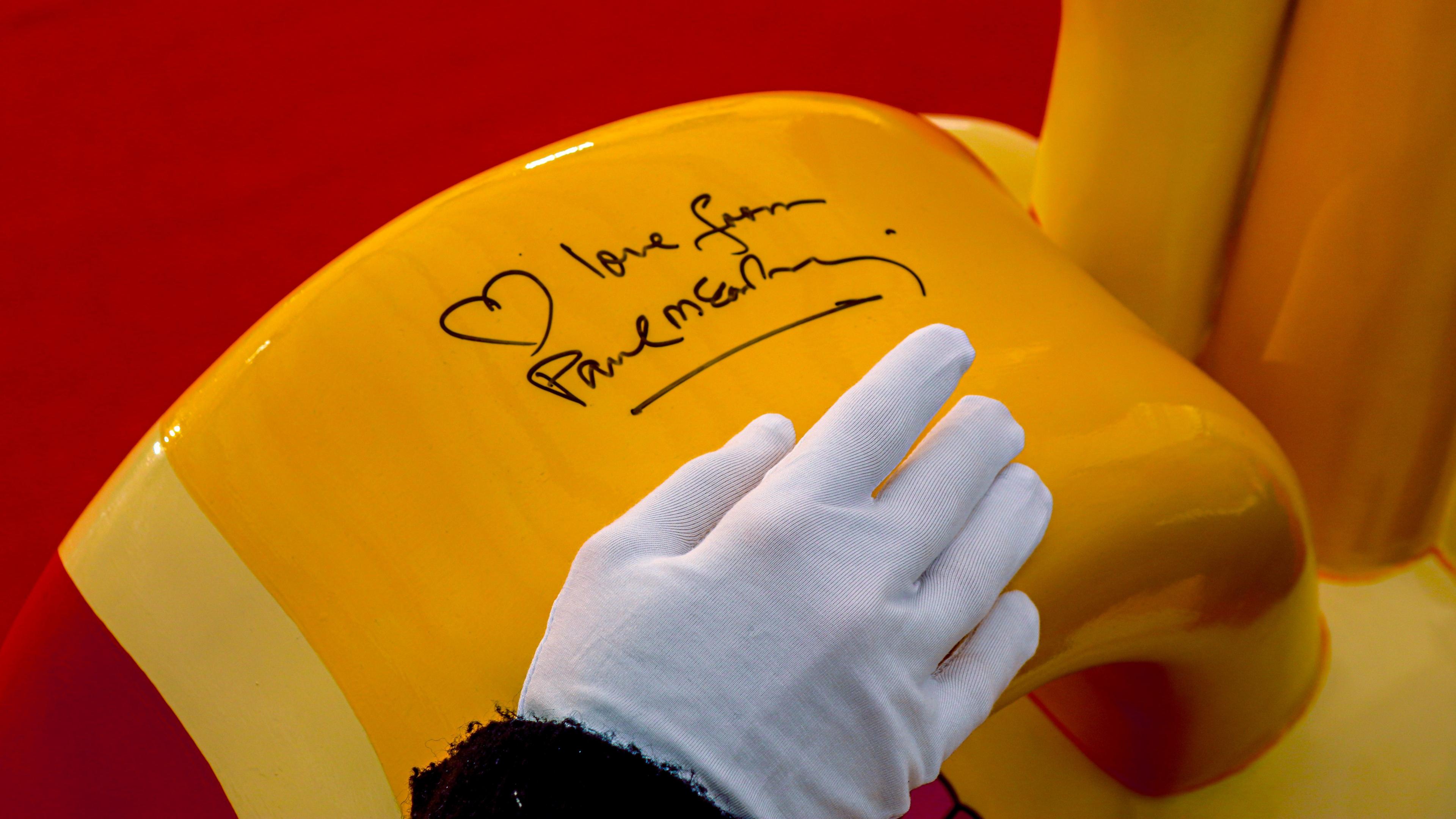 A close-up of an auctioneer's hand inside white glove. They are gently touching the top of the bright yellow Gromit sculpture, where Paul McCartney's signature is written alongside a heart in black permanent marker.