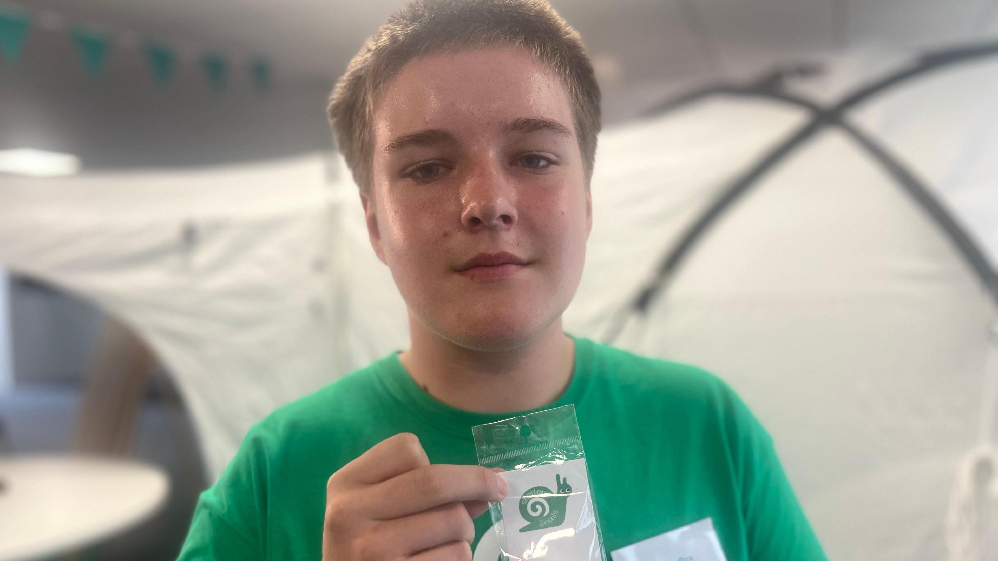 A teenager wearing a green T-shirt holds up a green snail. He is standing in front of a tent.