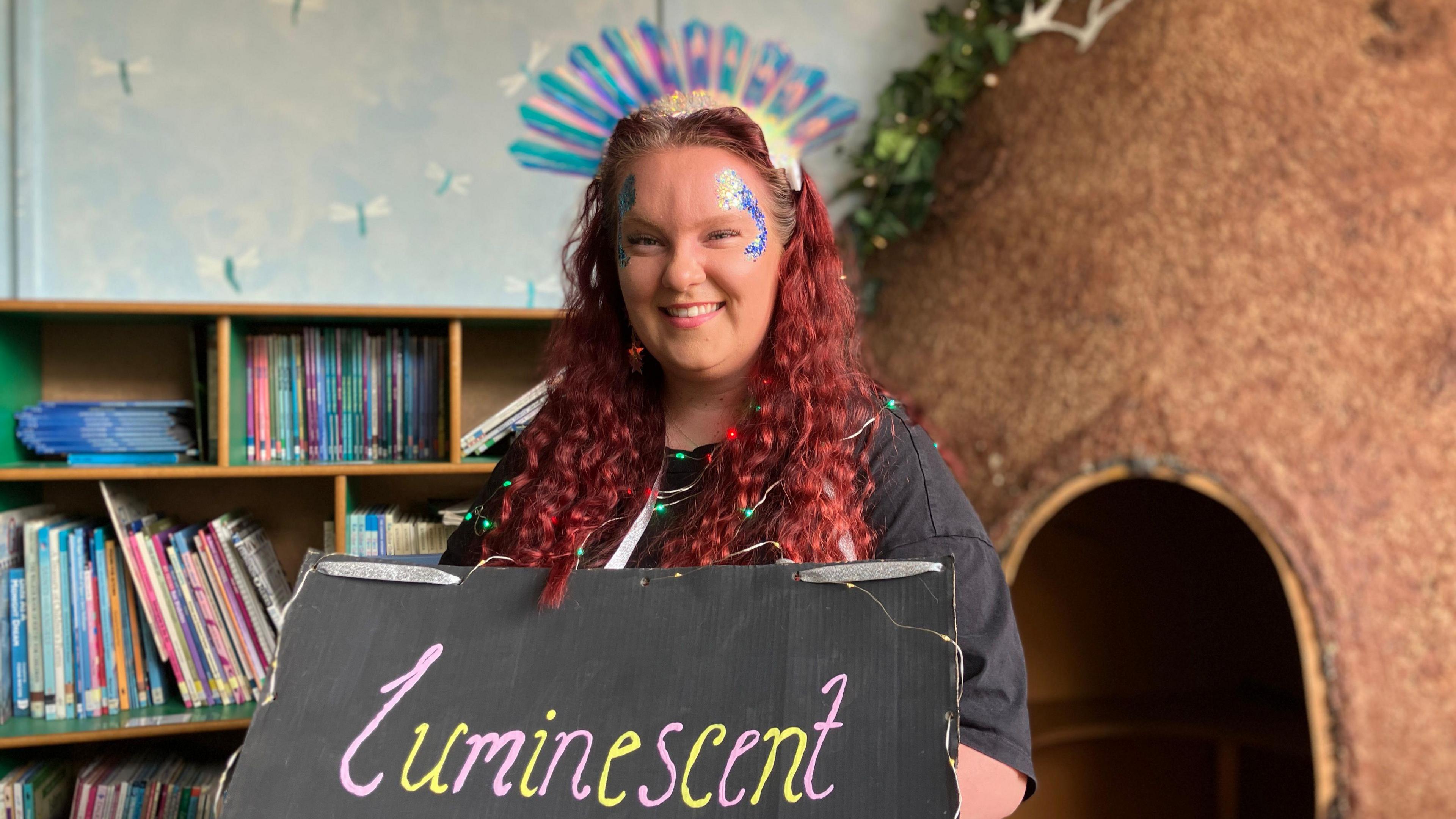 Teacher Lauren Noone with long red, curly hair and glittery make up. She is a holding a sign representing her word 'luminescent'