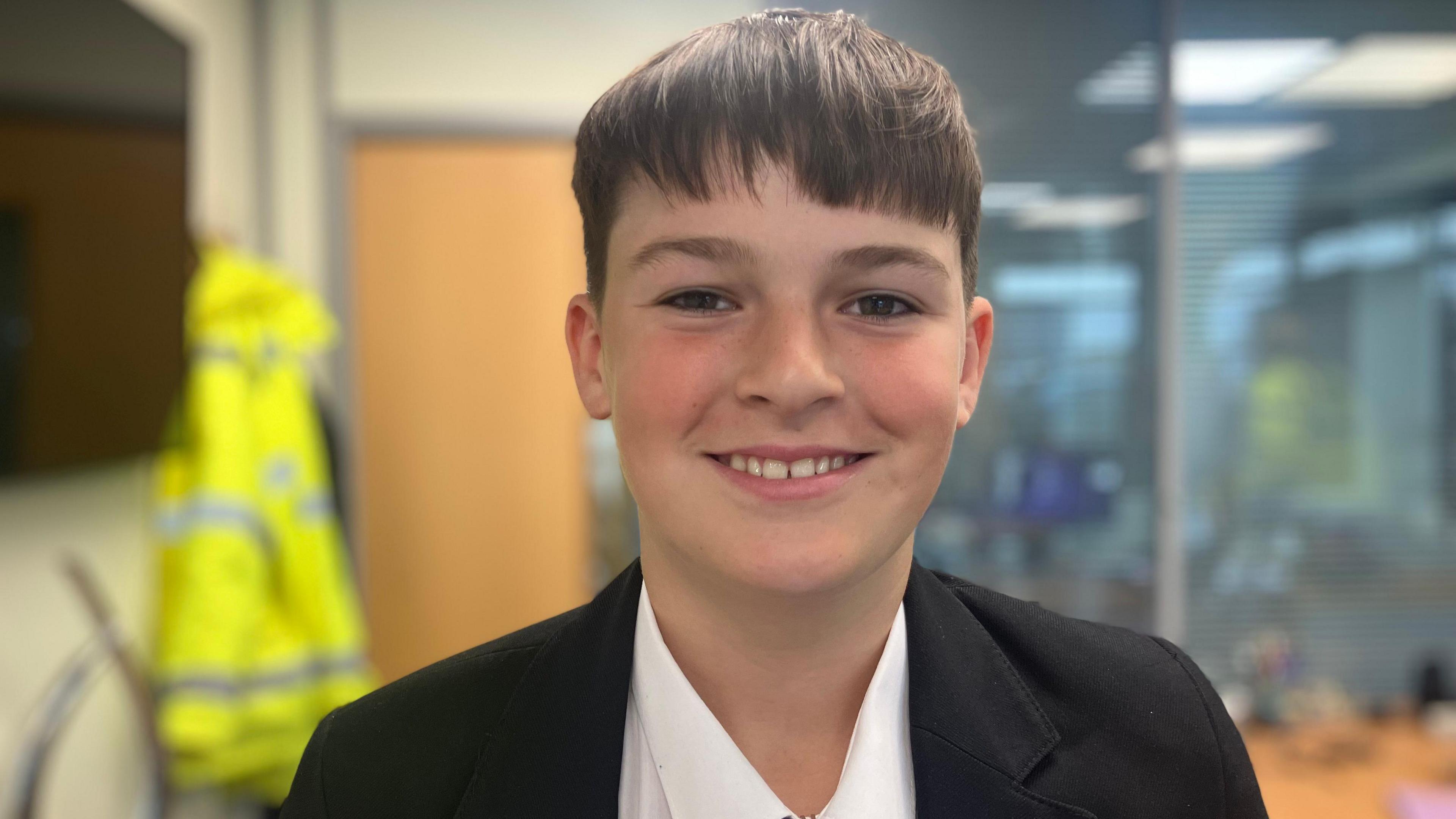 A boy with dark hair smiles at the camera. He wears a white shirt and a black blazer. Behind him a high vis hangs on a wall in the background.