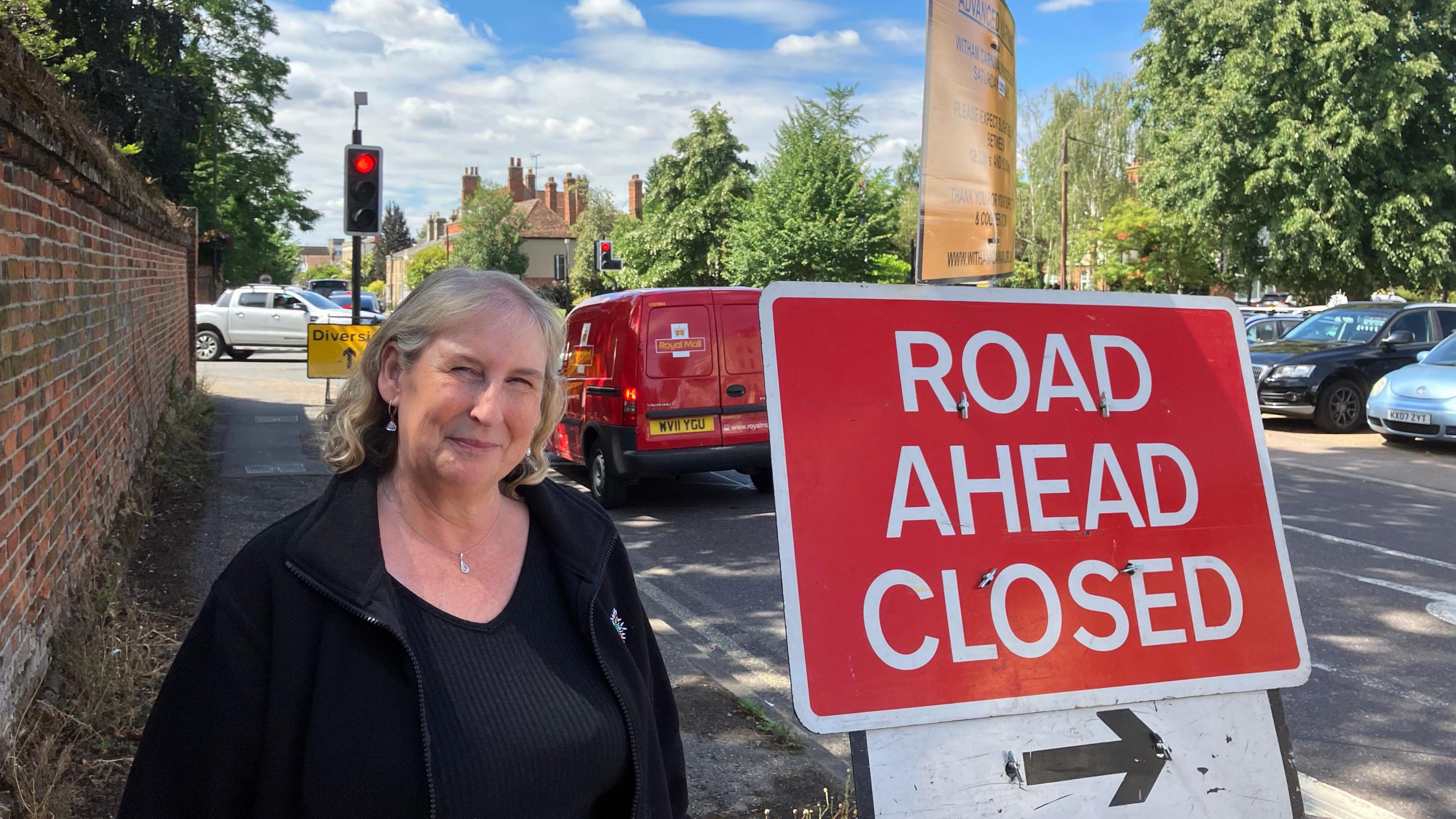 A woman standing next to a red "road ahead closed" sign, further in the background are some cars stopping at a red light