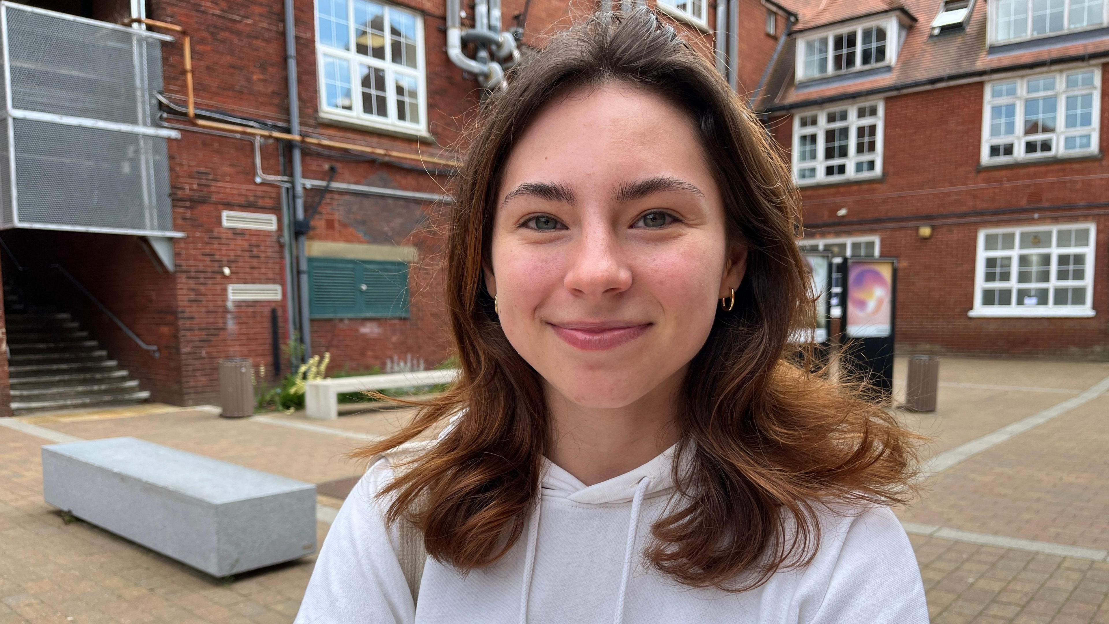 Izzie is 18 with brown wavy mid-length hair. She's smiling wearing a white hoodie.