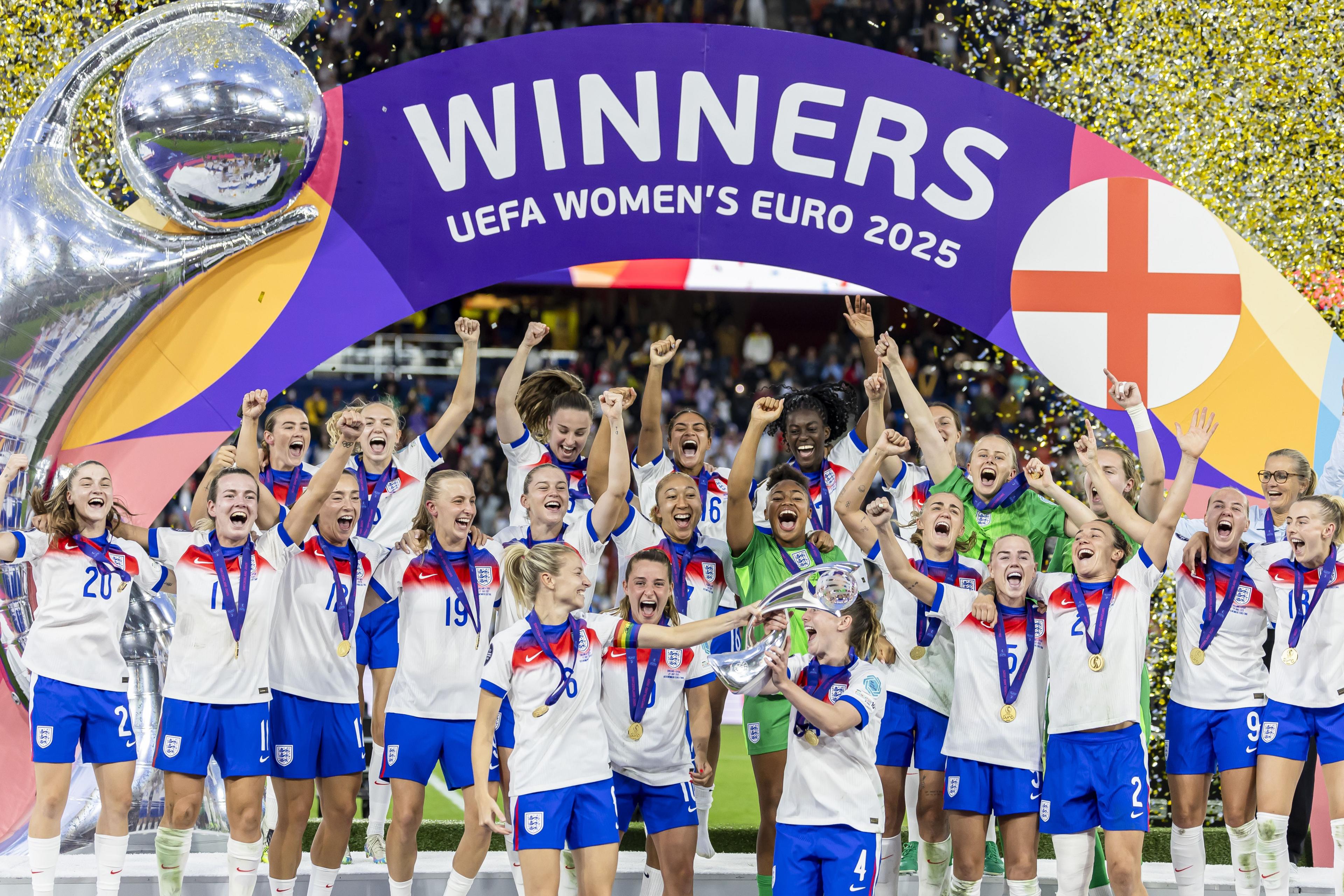 The Lionesses stand in front of a banner that says 'Winners Uefa Women's Euro 2025'. The women have fists in the air and are celebrating. There is confetti in the air.