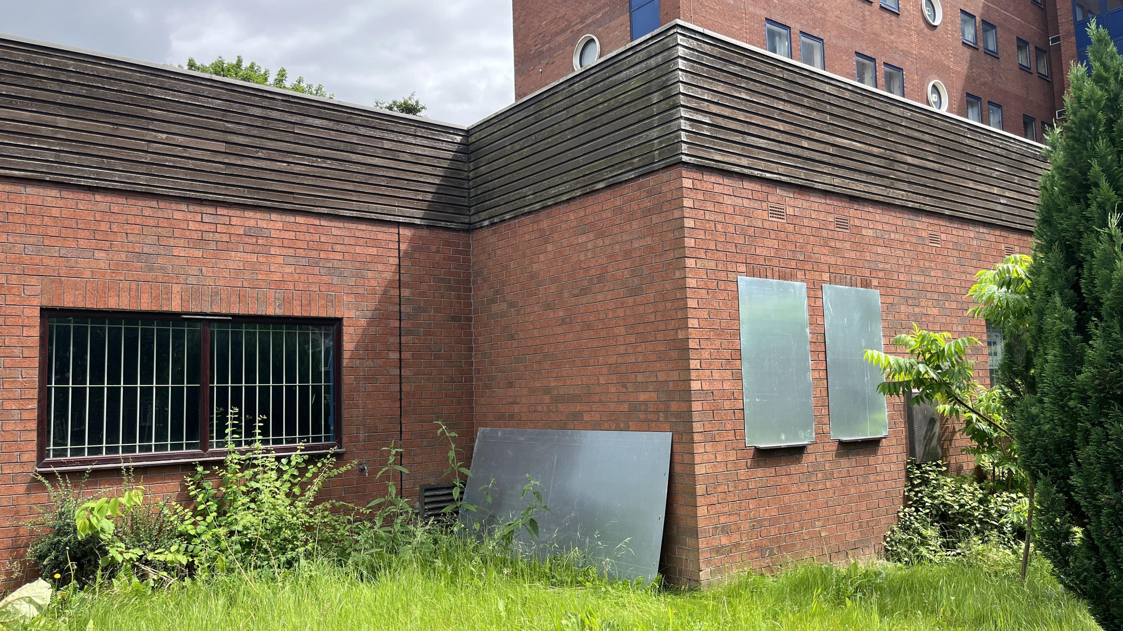 Overgrown grass and shrubs surround a red brick building. A window is visible and it has white bars behind it. A large metal sheet has been propped against a wall in the centre of the image and two other windows on the right have been boarded over.