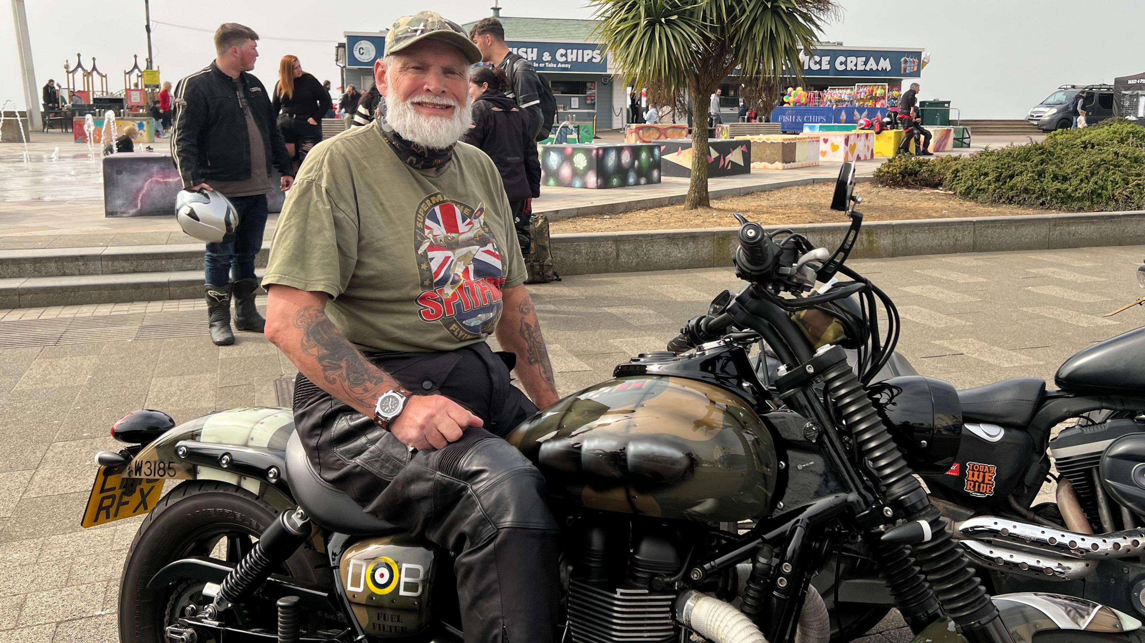 Paul Vincent sits on his motorbike. He wears a green cap with a green T-shirt and leather trousers. He has a long grey beard.