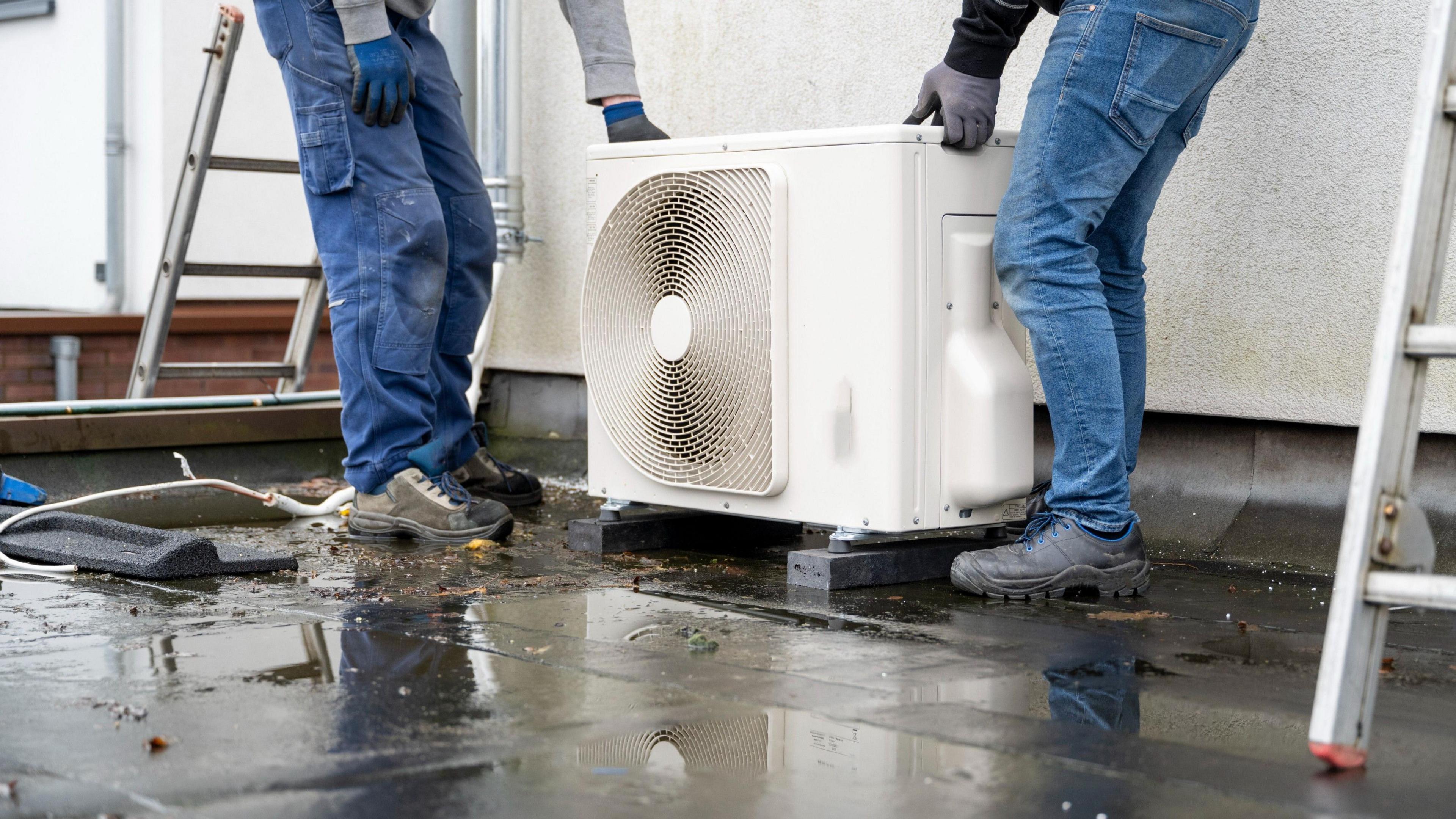 Two technicians installing an outdoor heat pump unit on a wet surface