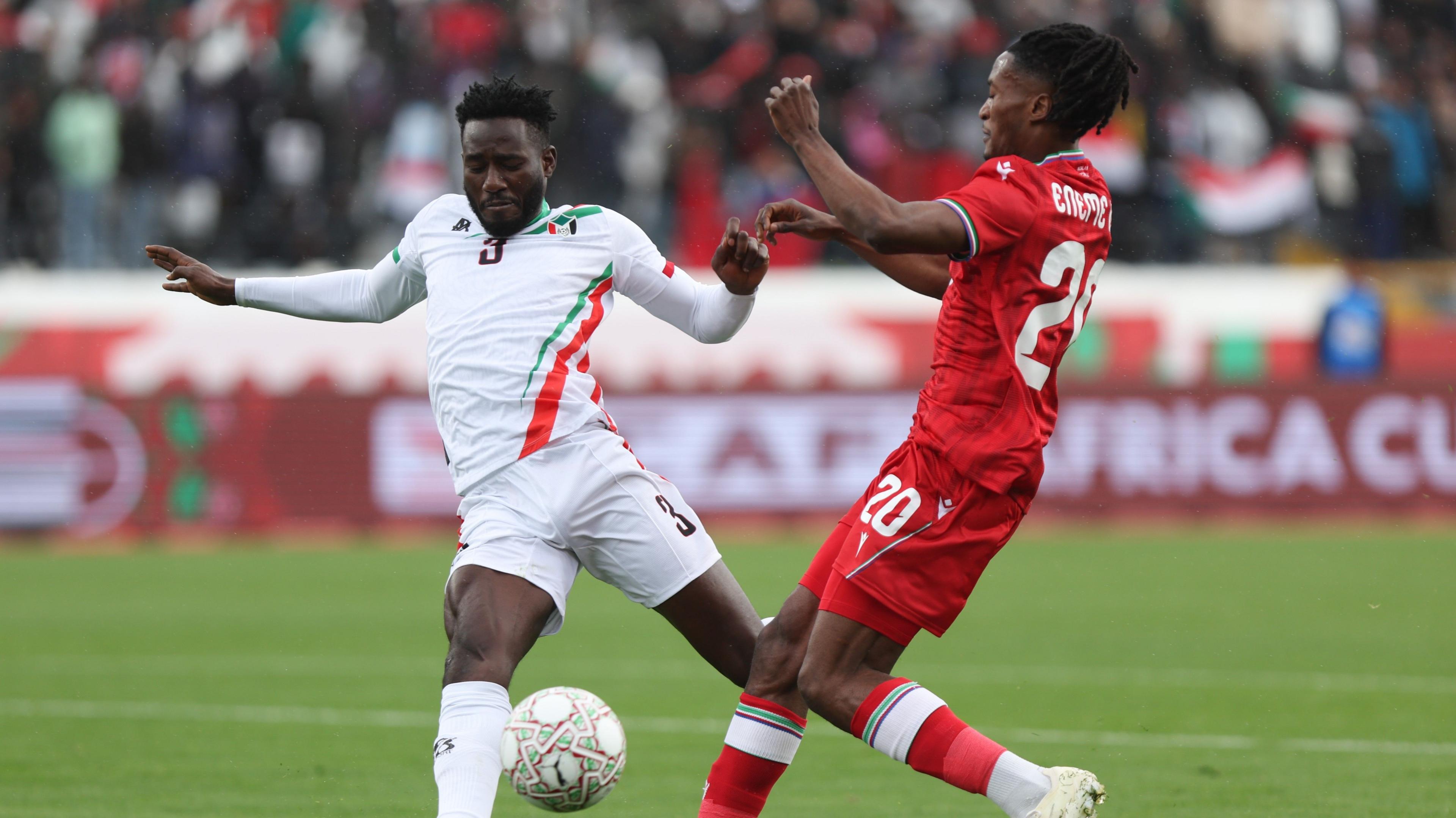 Footballer Mohamed Ering is pictured playing football for the Sudan national team at the Africa Cup of Nations 2025, challenging for a football in a white kit with green and red details and the number 3 in black on the chest and shorts. Ering is challenged by Equatorial Guinea footballer Santiago Eneme, wearing a red kit with white trim and his name and number 20 on the shirt and shorts, as well as white boots
