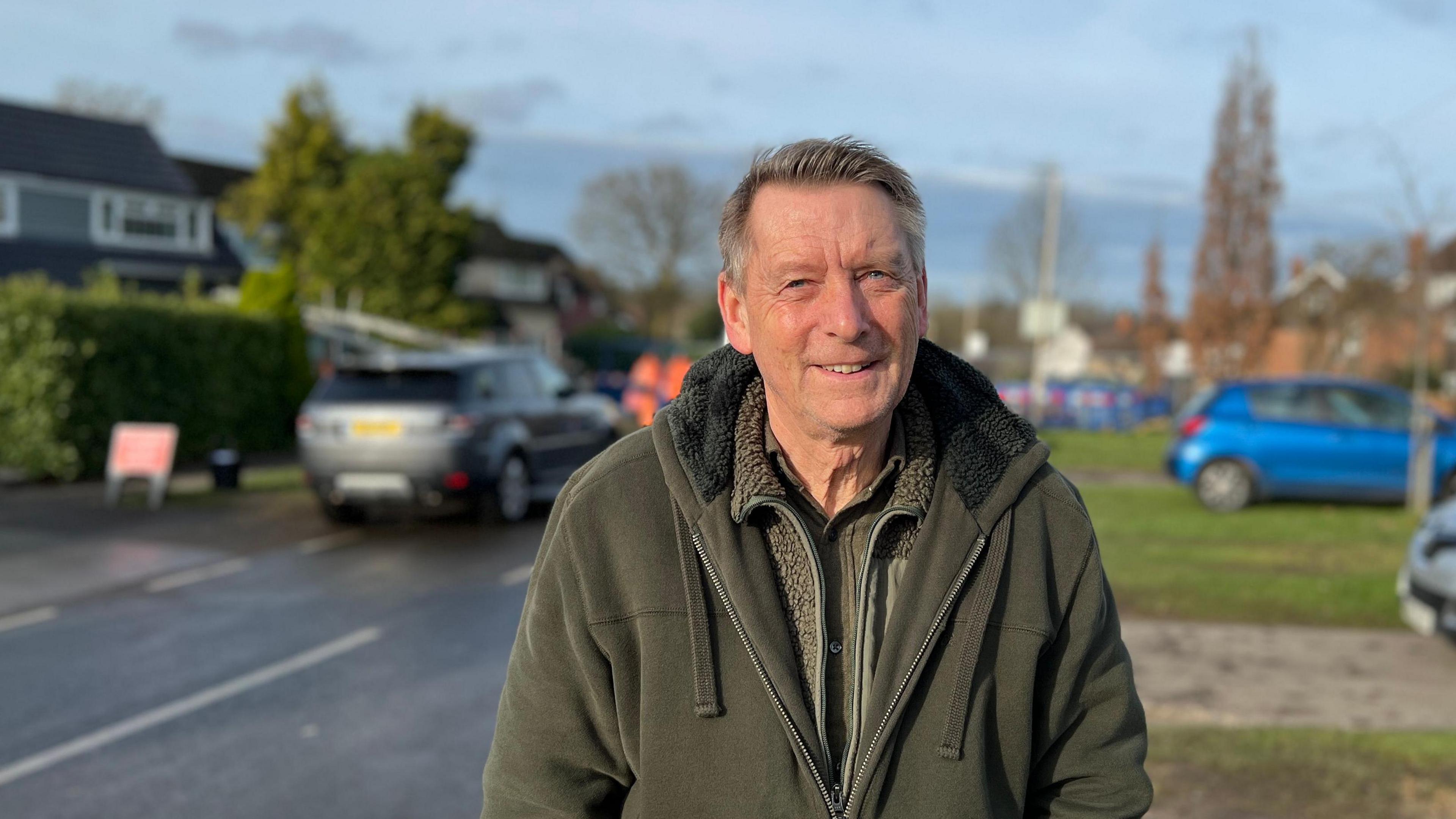 Terry Polden is smiling at the camera and wearing a green fleece. He is standing on the road with the burst water main in the background. 