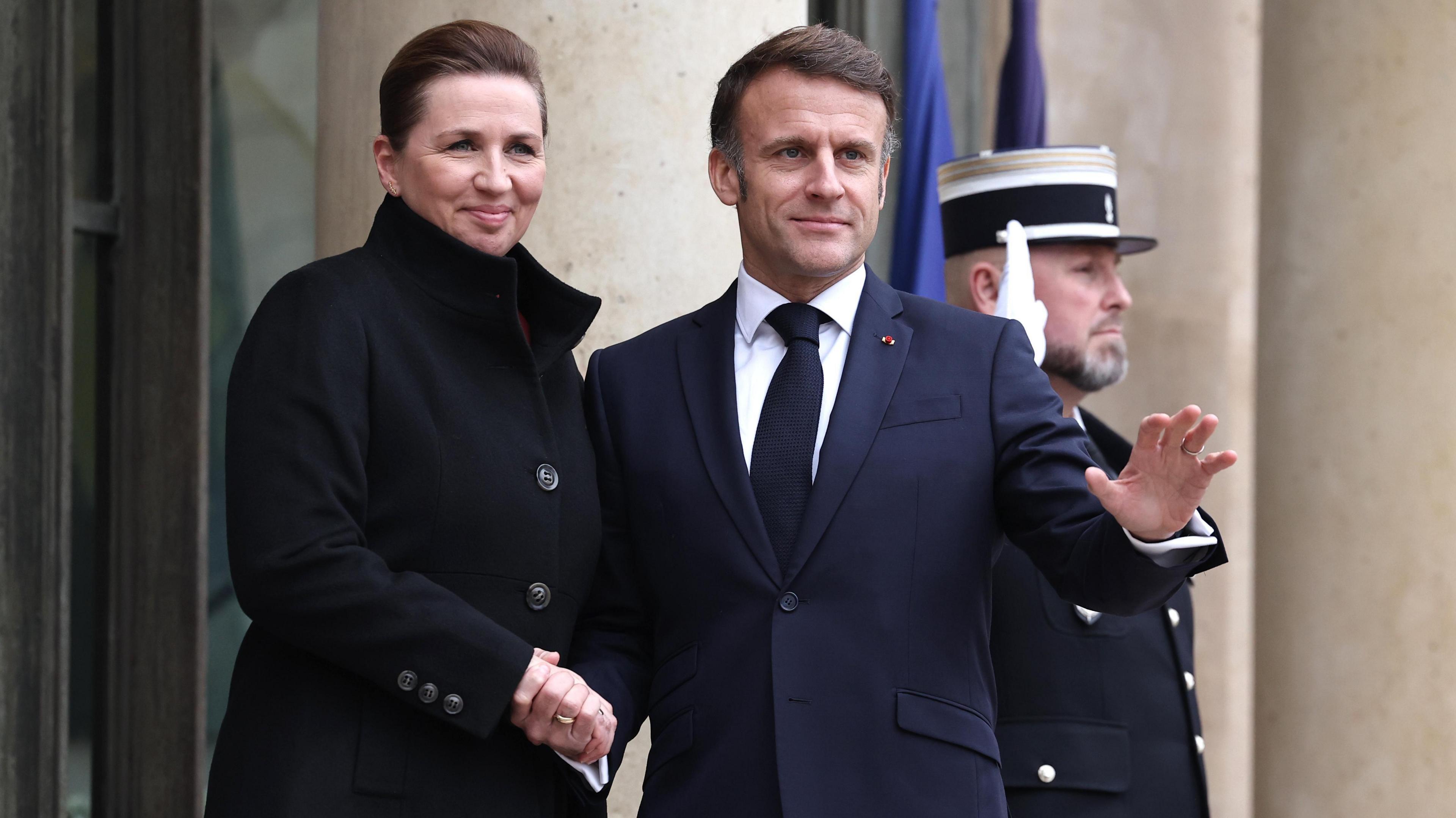 Mette Frederiksen, wearing a dark coat, shakes President Macron's hand. He is wearing a dark blue suit jacket and a tie.