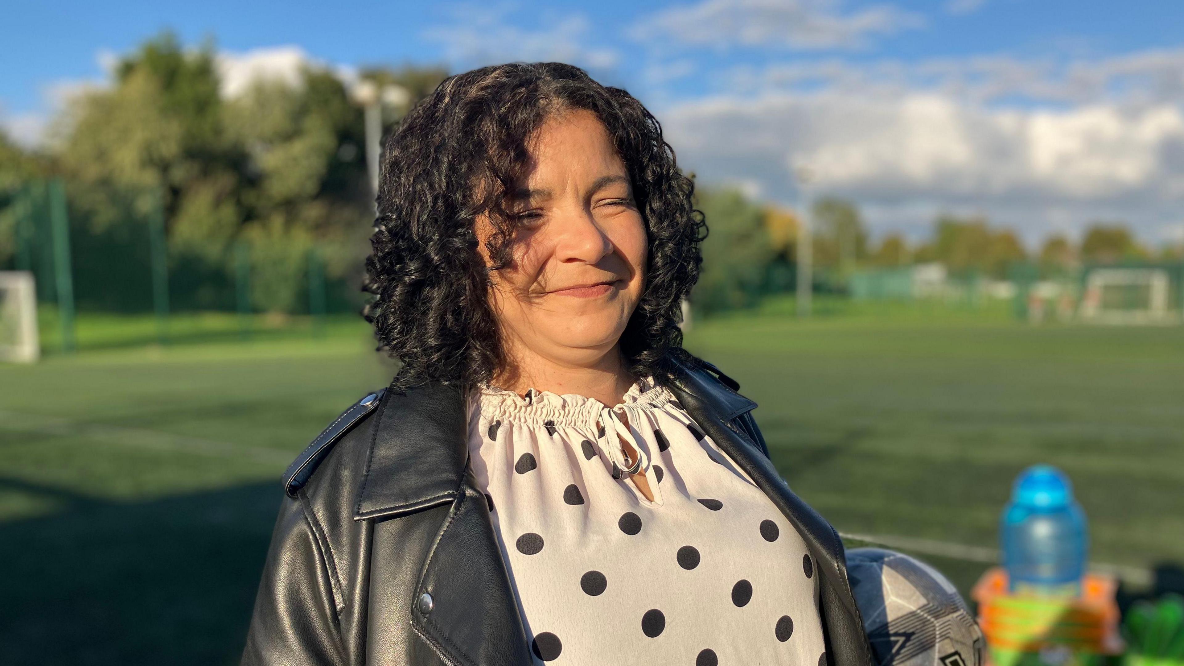 A lady with dark curly hair, a white and black polka dot top and a black leather jacket. She is smiling and holding a ball. 