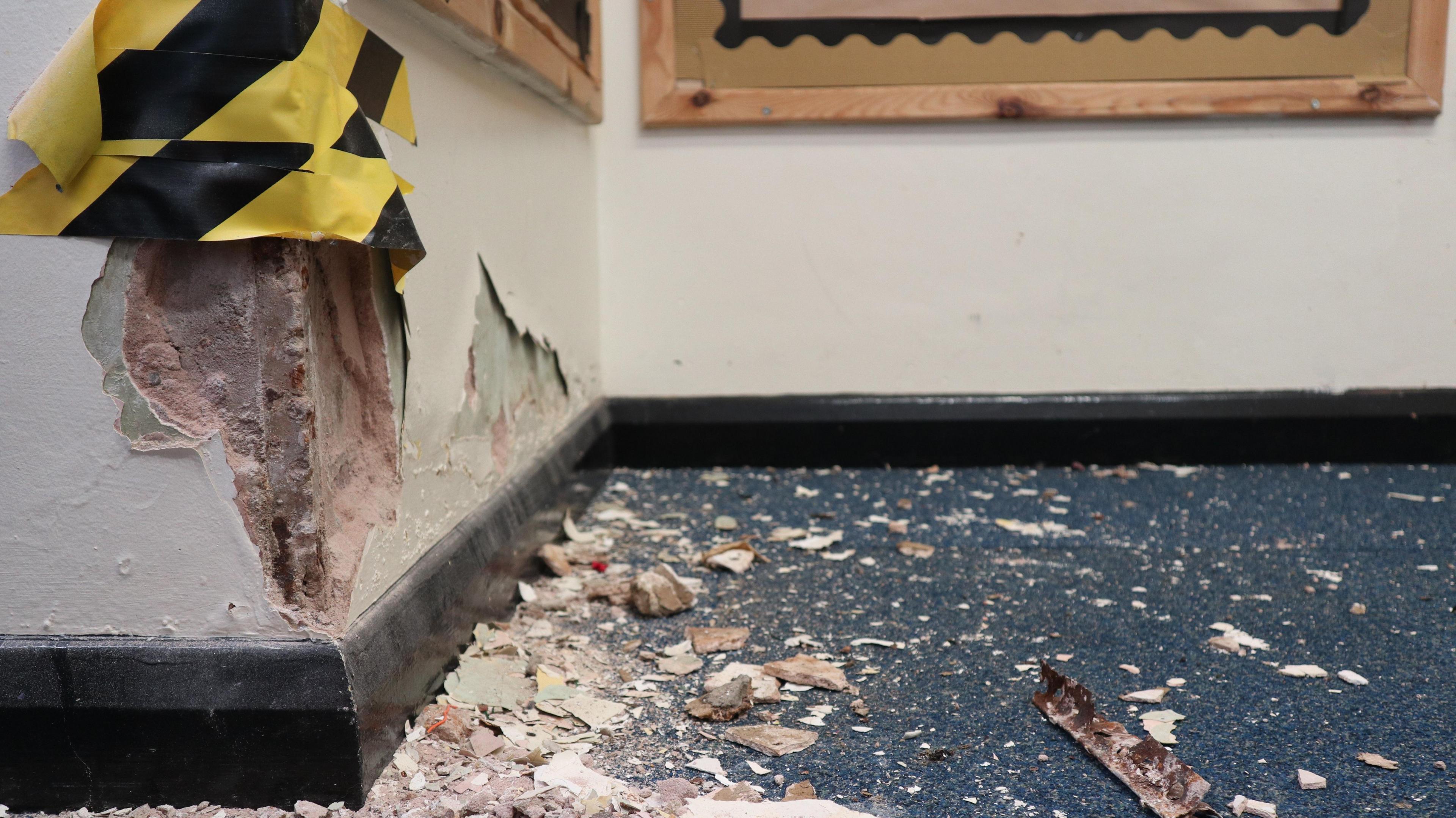 The image is a close up shot of the corner of a classroom. Wall displays can be seen in the background. In the foreground the blue carpet is covered in bits of plaster, and there is a deep hole in the corner of the wall above it, which has three bits of yellow warning tape on it.