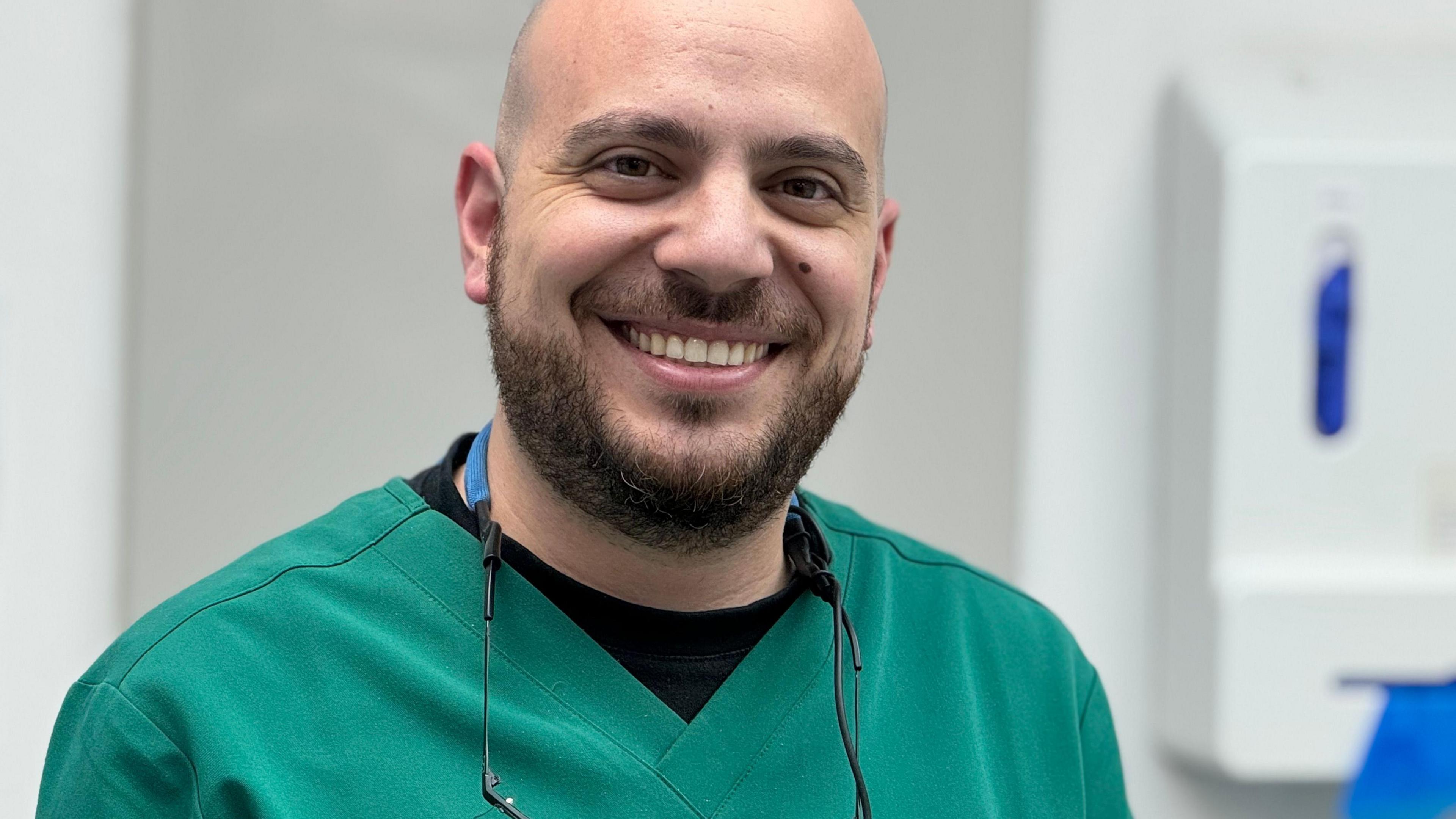 A shaven-headed man smiles into the camera. He has a short dark beard and wears a dark green top. A necklace runs around his neck and hangs beyond the bottom of the frame.