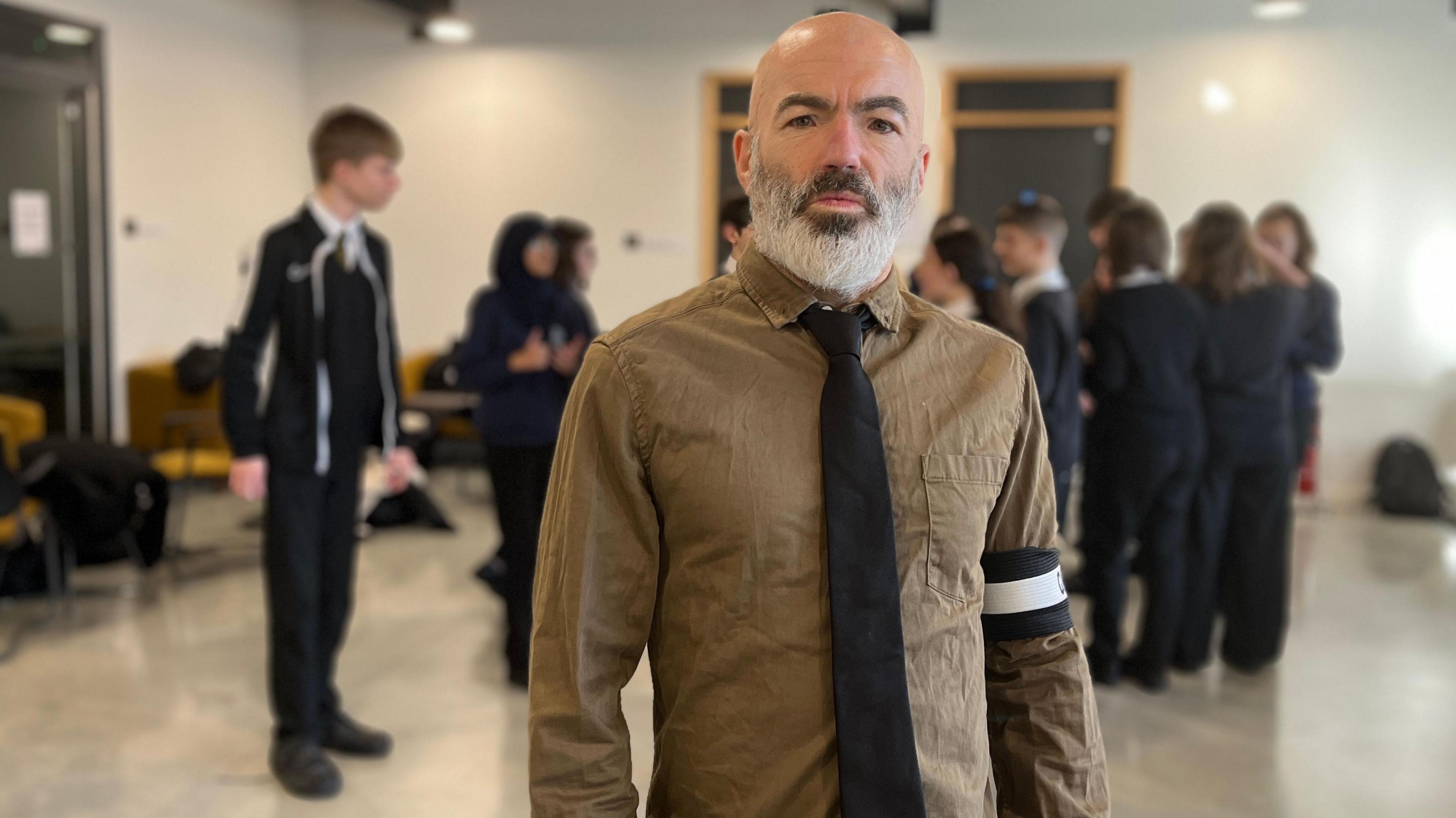 A man with a shirt and tie and armband stands in front of a group of school students