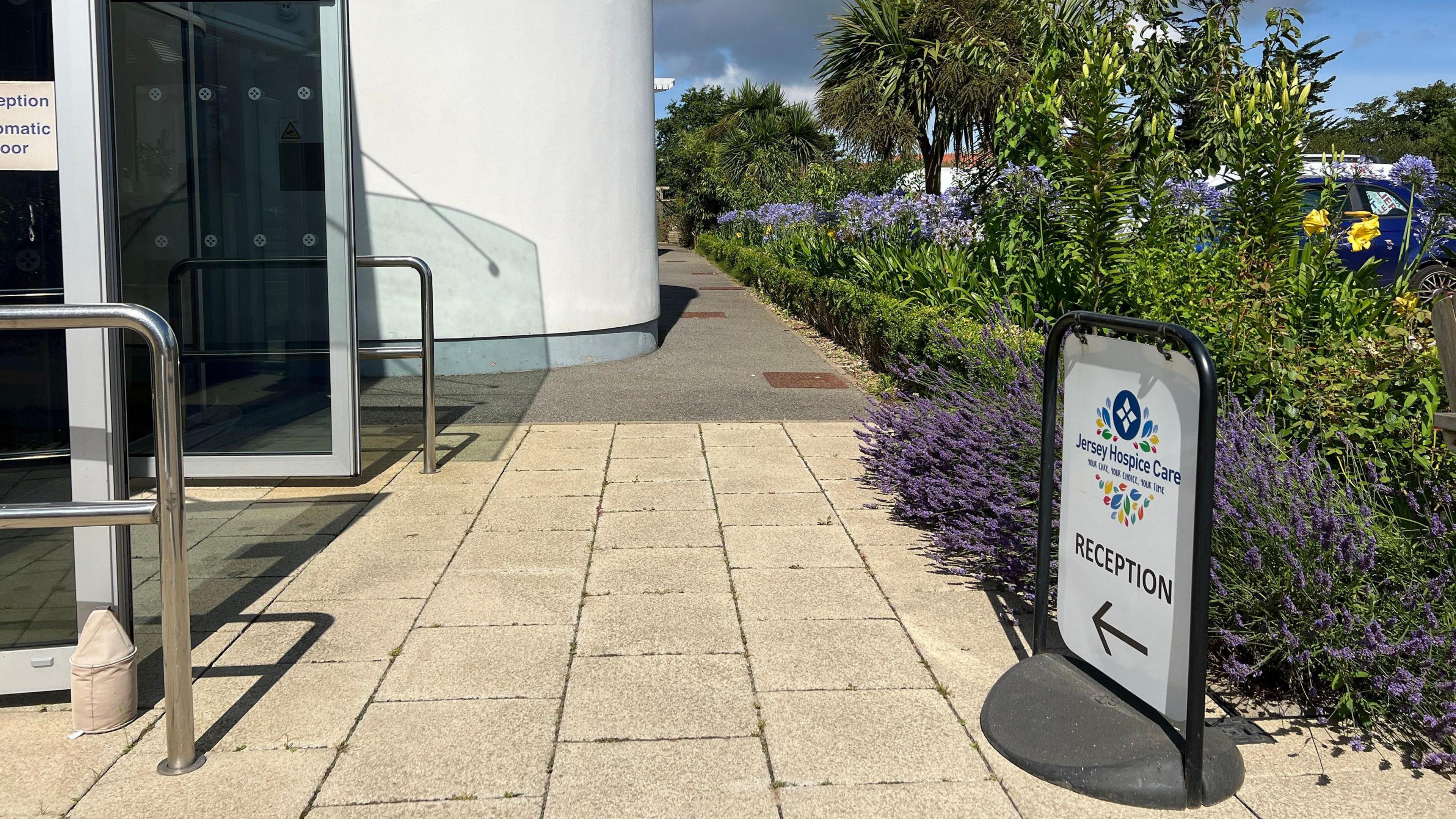 Entrance to a hospice care facility with an automatic glass door. A pathway bordered by greenery and purple flowers leads to the entrance. A 'Reception' sign with an arrow points toward the door, and another sign reads 'Hospice Care.' The sky is partly cloudy with blue patches