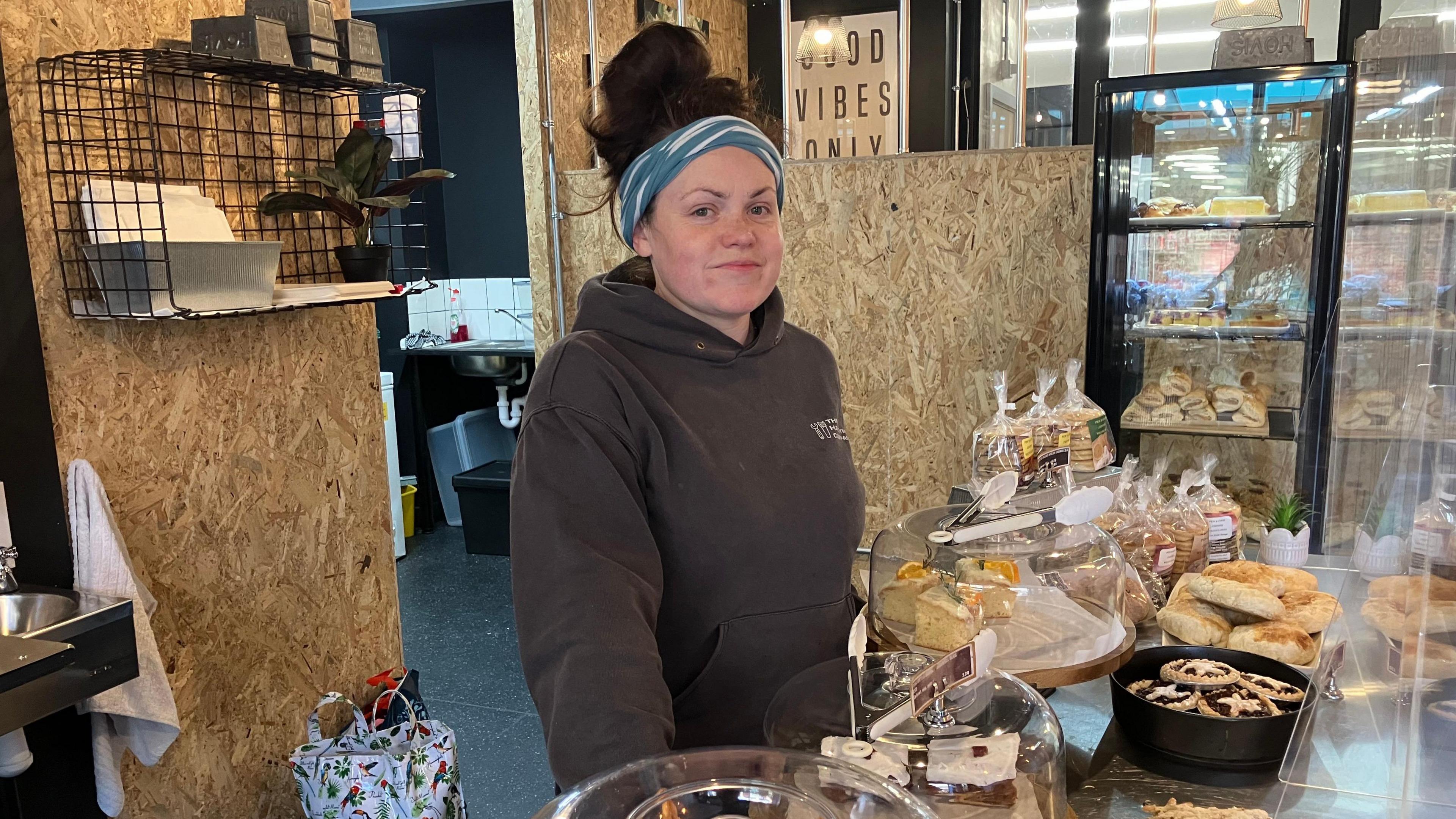 Amelia Jones from The Little Welsh Bakery is standing behind the counter looking into the camera and in front of her are some baked goods. She is wearing a black hoodie and has dark hair tied back in a ponytail, with a blue-and-white headband. 