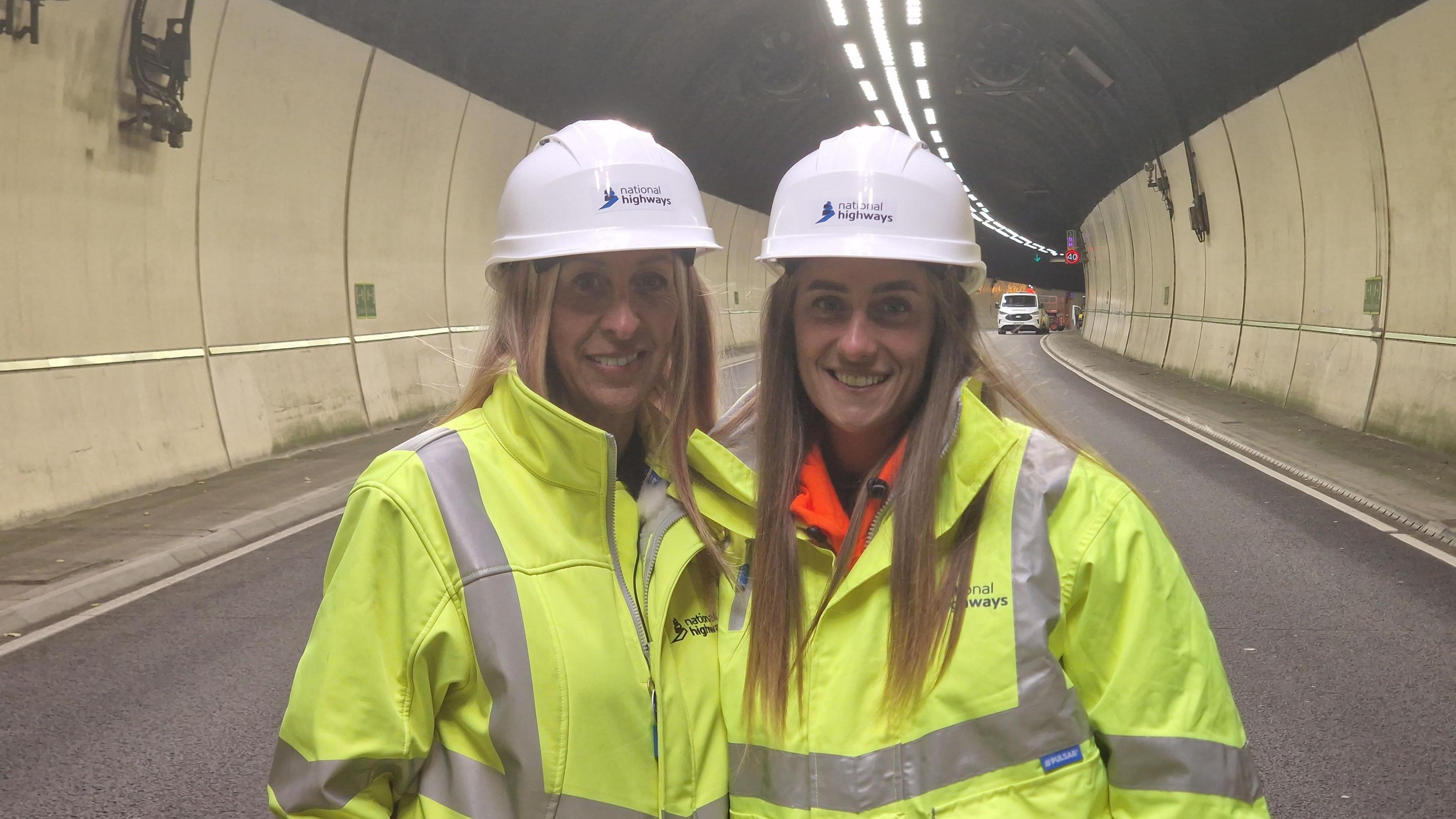 A mother and daughter both with long blonde hair stood in the centre of a dual carriageway in a tunnel. The road is clear of vehicles apart from a white van in the background on the right. The woman are wearing yellow high-vis jackets and white hard hats while smiling at the camera