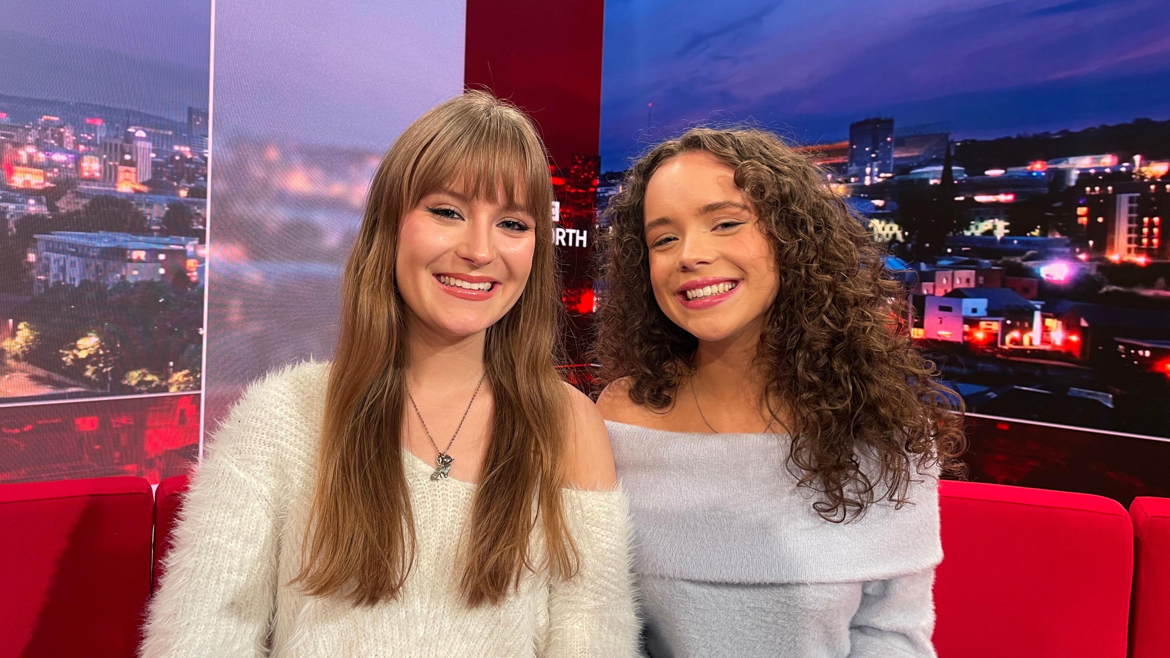 A close-up of Grace (left) and Hannah (right), who are looking directly into the camera. Grace is wearing a fluffy white jumper and has long, straight, light brown hair. Hannah is wearing a fluffy, light blue jumper and has long, curly, dark brown hair. 