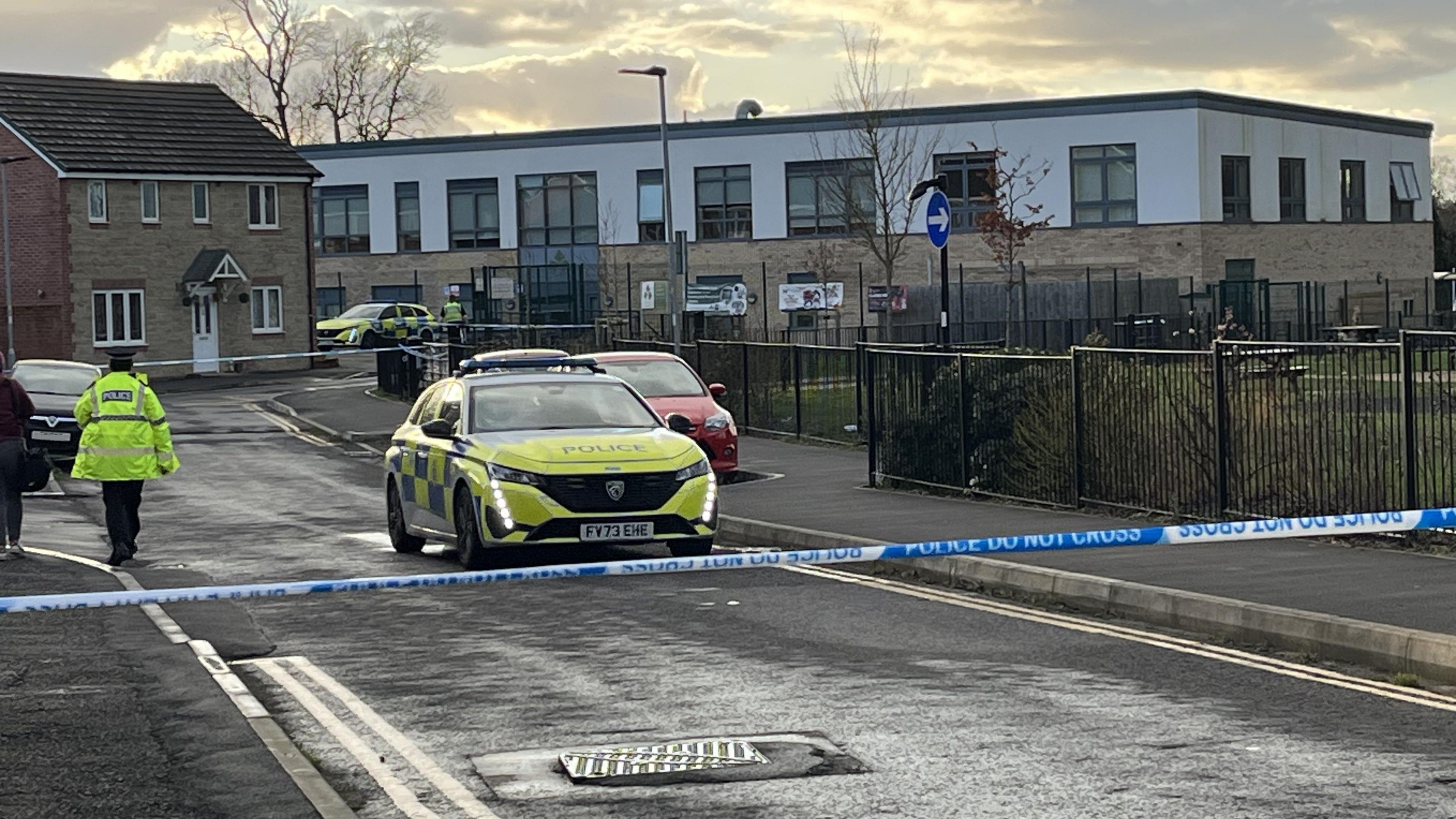 In the foreground, there is blue and white police tape that says "do not cross". In the background, a school, a police car and a police officer with his back to the camera in their hi-vis yellow uniform 