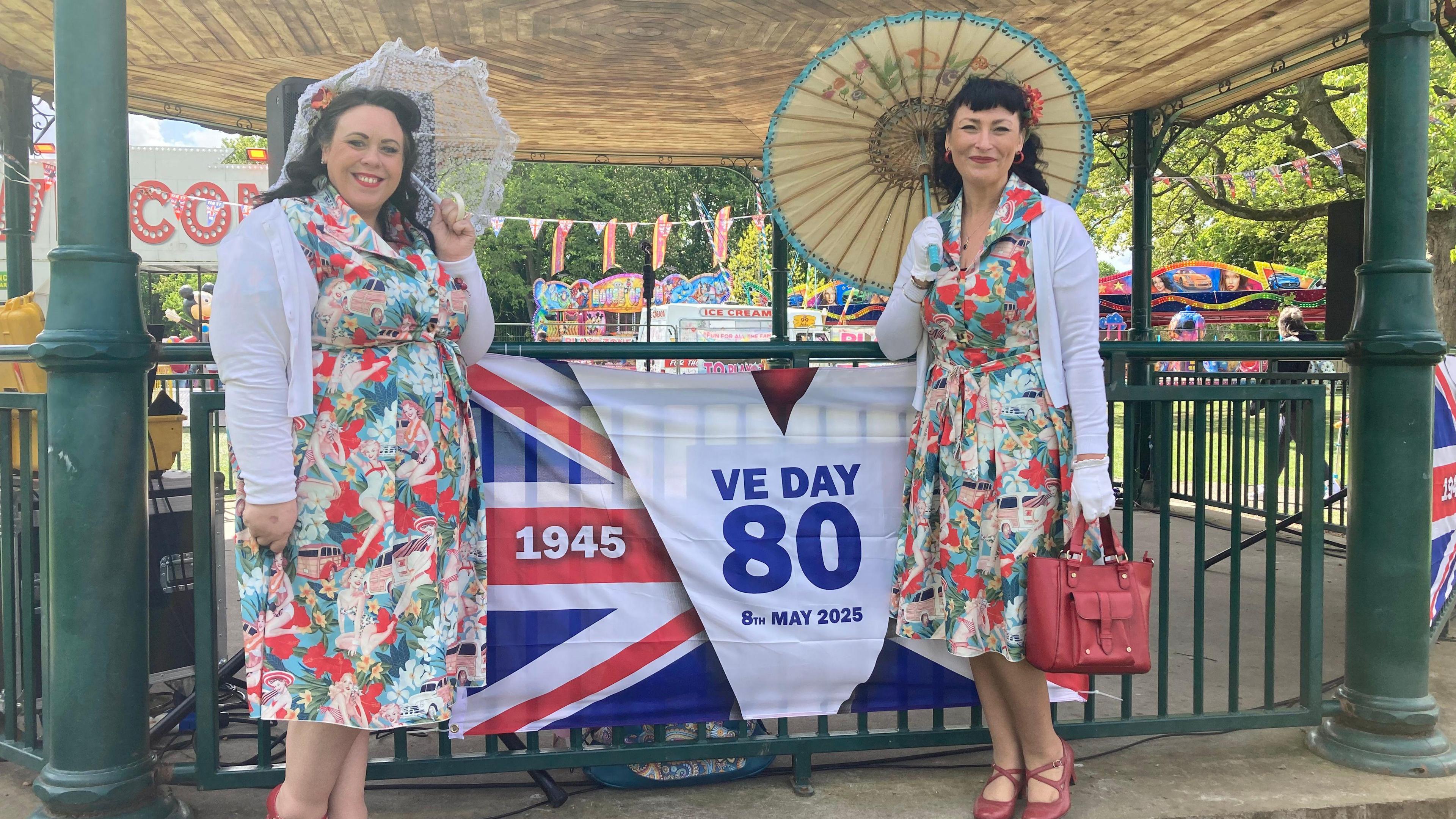 Two women dressed in vintage floral dresses and white cardigans stood with parasols beside a VE Day 80th anniversary banner. They stand in front of a bandstand at a lively outdoor celebration, with fairground rides and Union Jack bunting in the background.