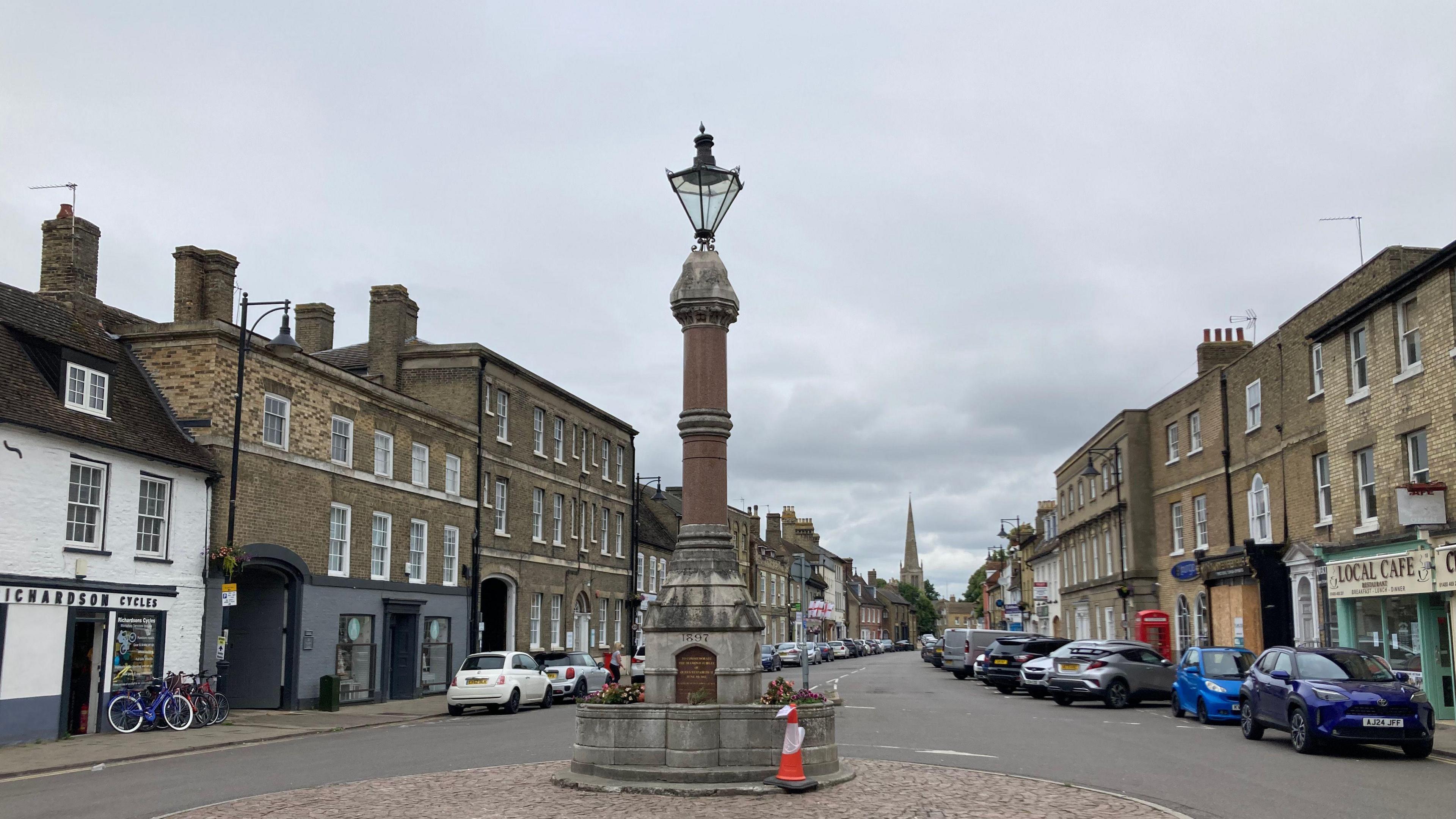 The broadway in St Ives. View of the Queen Victoria memorial with a large lamp on top, and a street with parked cars and Georgian buildings either side. There are shops including a cycle shop and local cafe.