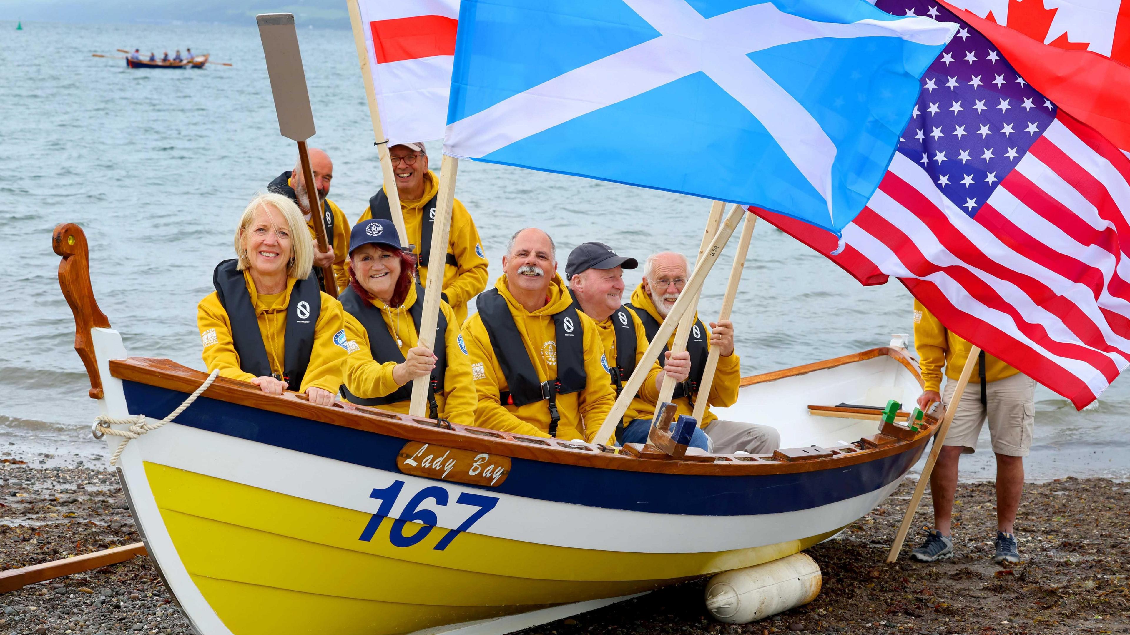 A boat crew dressed in bright yellow wave a range of flags while sitting in a yellow, white and blue boat on the south of Scotland shoreline