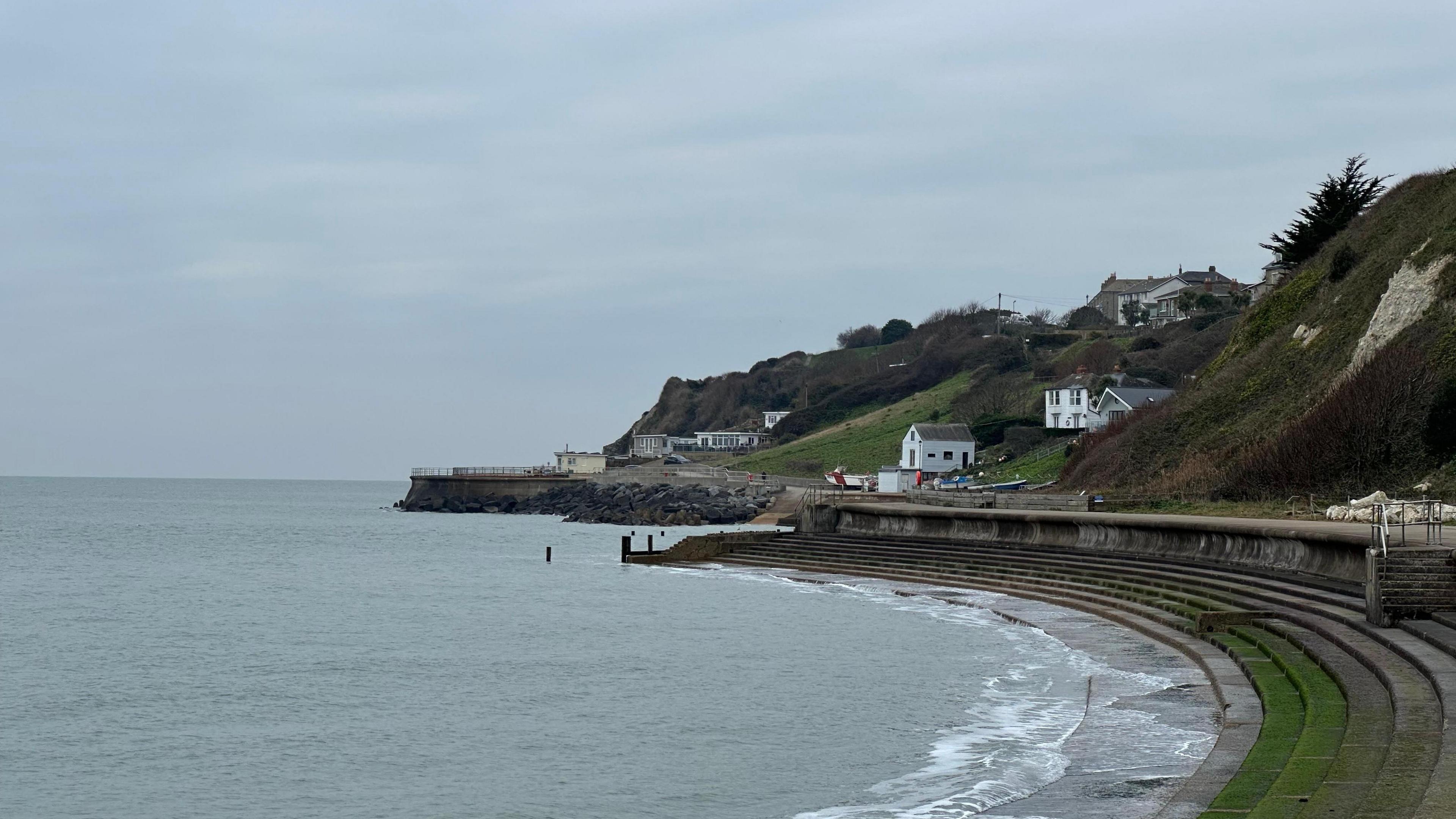 A view along the coast at Ventnor on the Isle of Wight. The sea wall has steps down to the sea. The cliffs rise above the wall with several white buildings. The cliffs have tree and grass cover. The sky overhead is grey. 