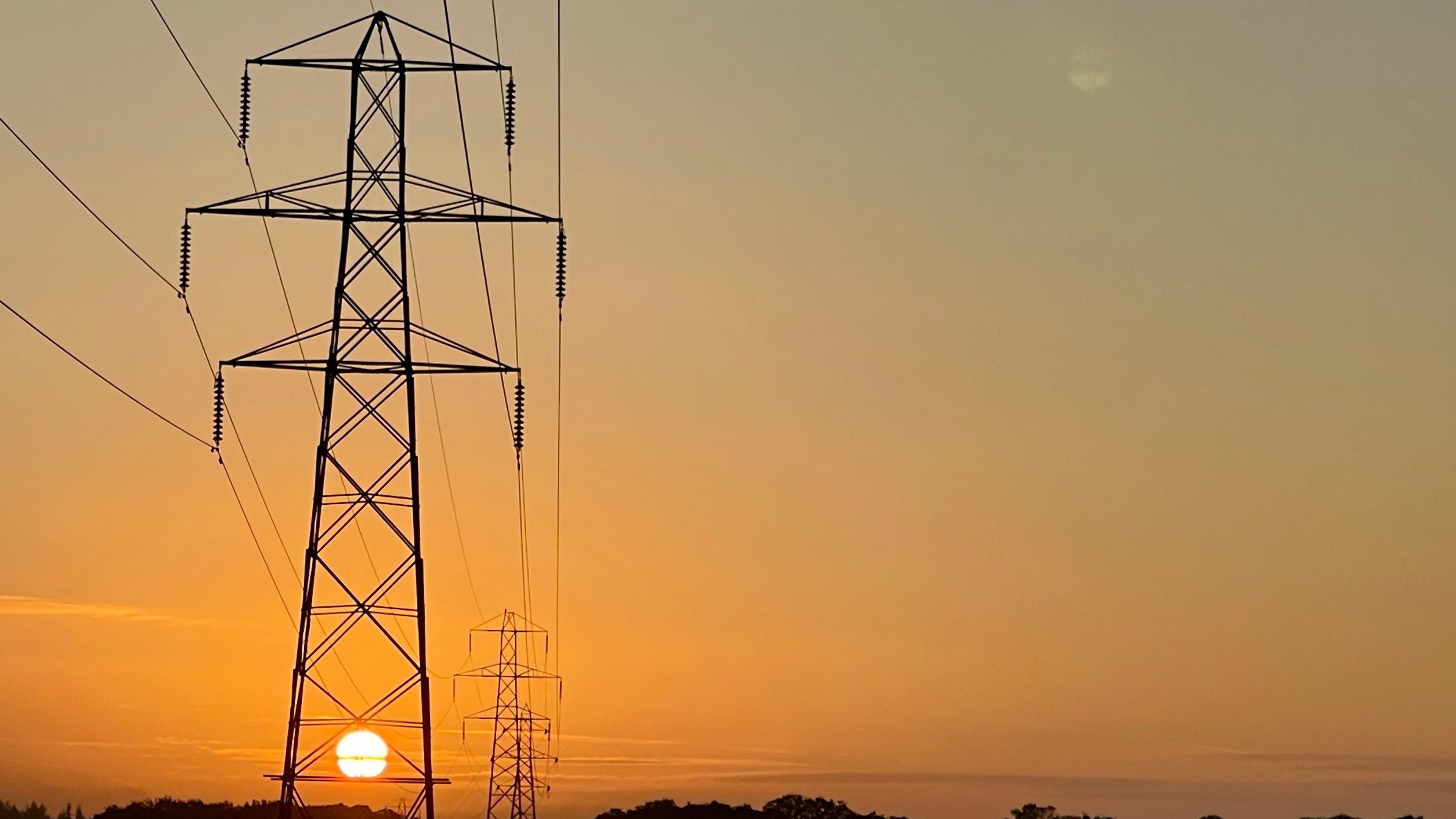 The rising sun shines orange through the metalwork of an electricity pylon which rise above the silhouette of trees on a low horizon. The wide expanse of sky is illuminated with a graduating orange emanating from the early morning sun.