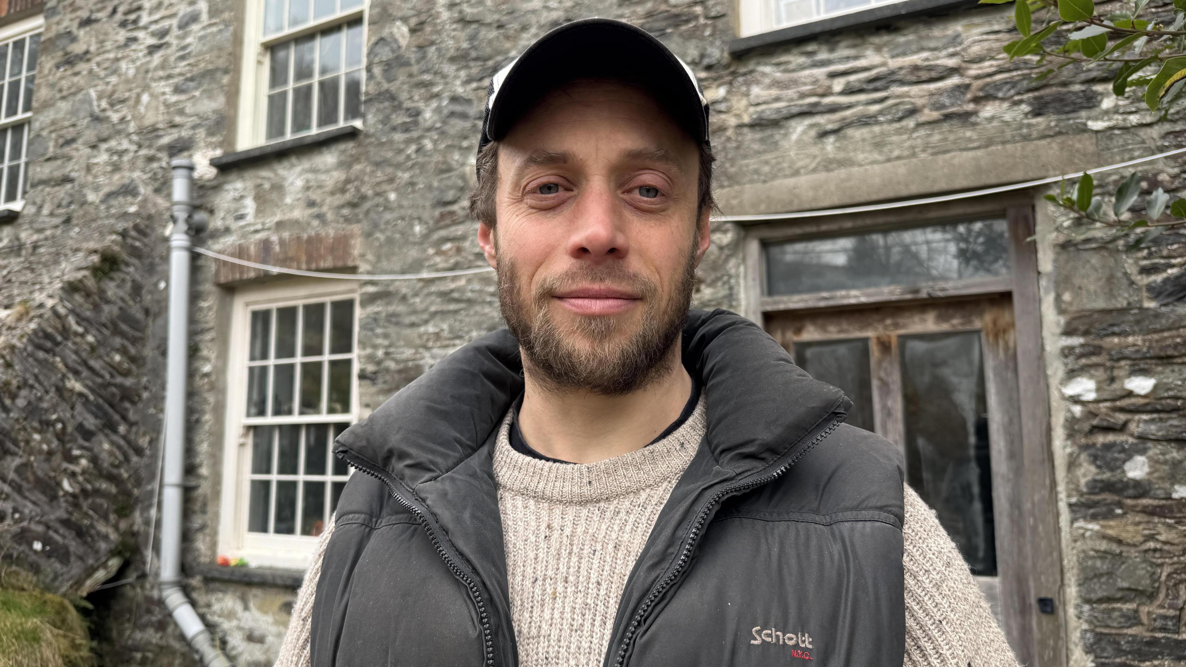 Owen Shiers outside the house he rents as part of a co-operative in Eglwys Fach, Ceredigion. He is wearing a baseball cap, a beige knitted jumper, and a black bodywarmer on top. He has a short, light brown beard. The grey stone building behind him has a wooden door and a white window sill. 