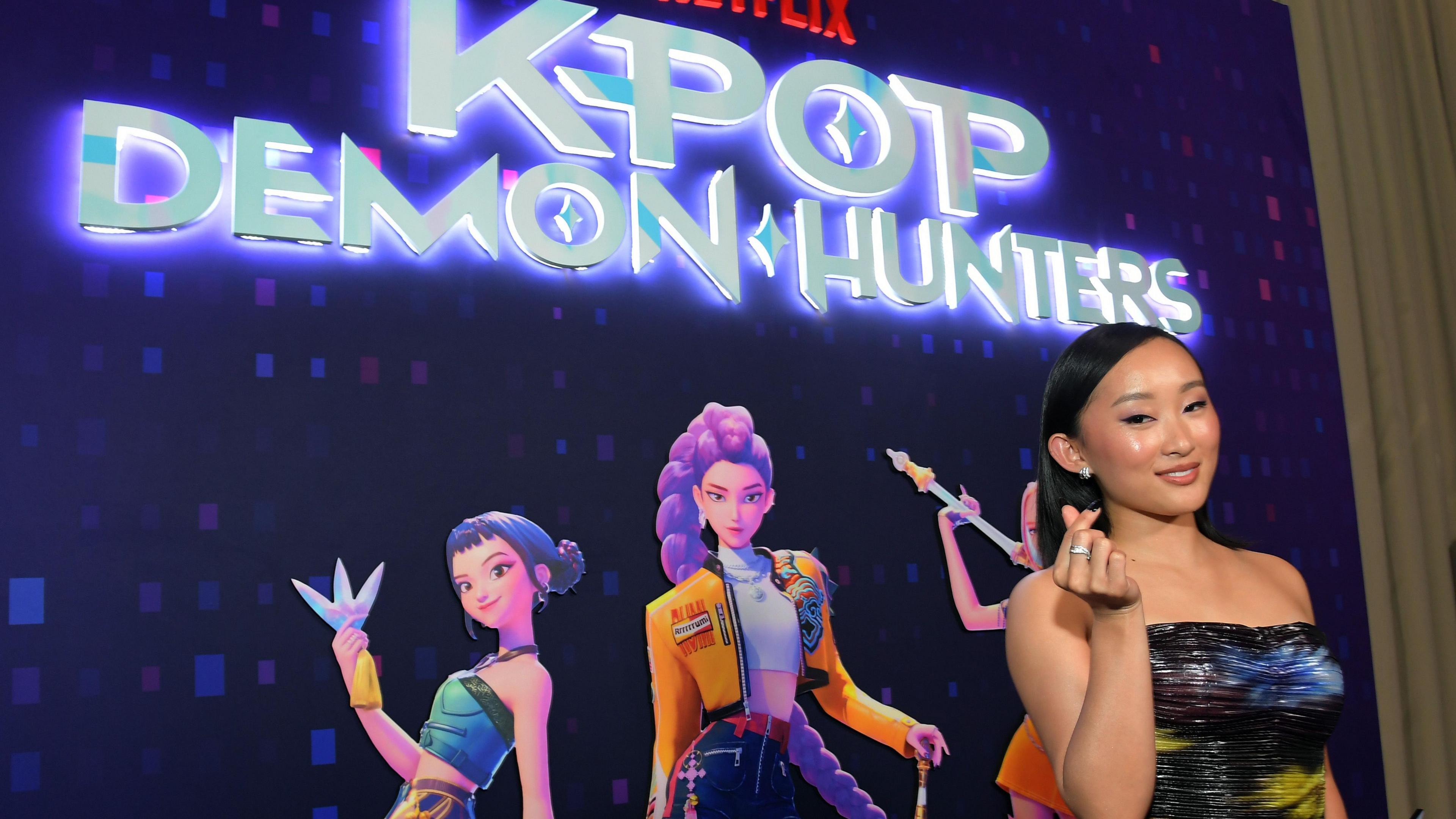 SunHee Seo poses in front of a poster for KPop Demon Hunters - the film's logo is lit up with a a purple glow and the three main characters pull poses beneath it on a purple background. Sunhee wears a shoulderless gown and appears to be clicking her fingers as she smiles.