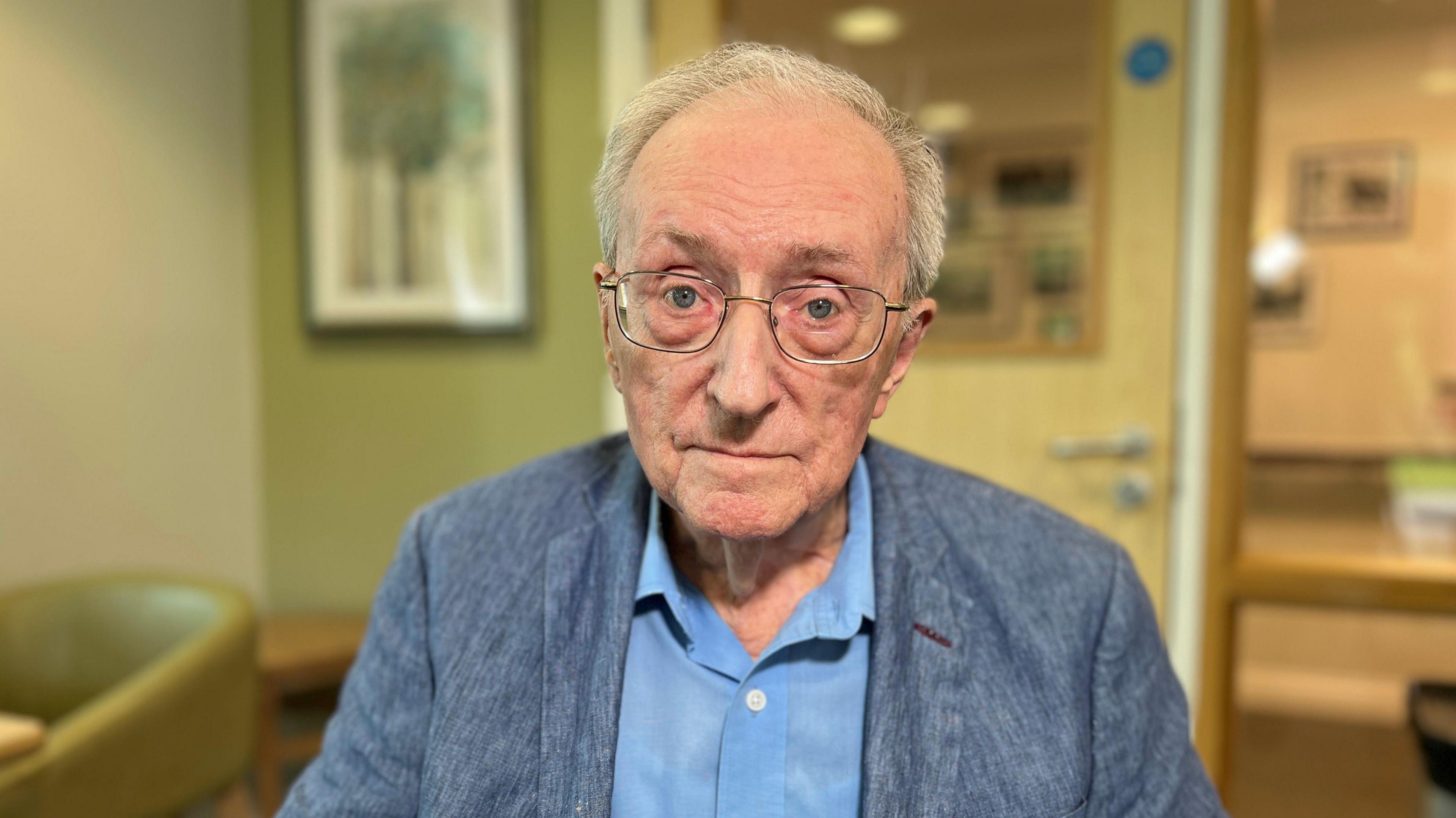 An older man sits in a room resembling a waiting area, wearing a blue shirt and jacket. The background includes chairs, a framed picture on the wall, and a door with glass panels leading to another room