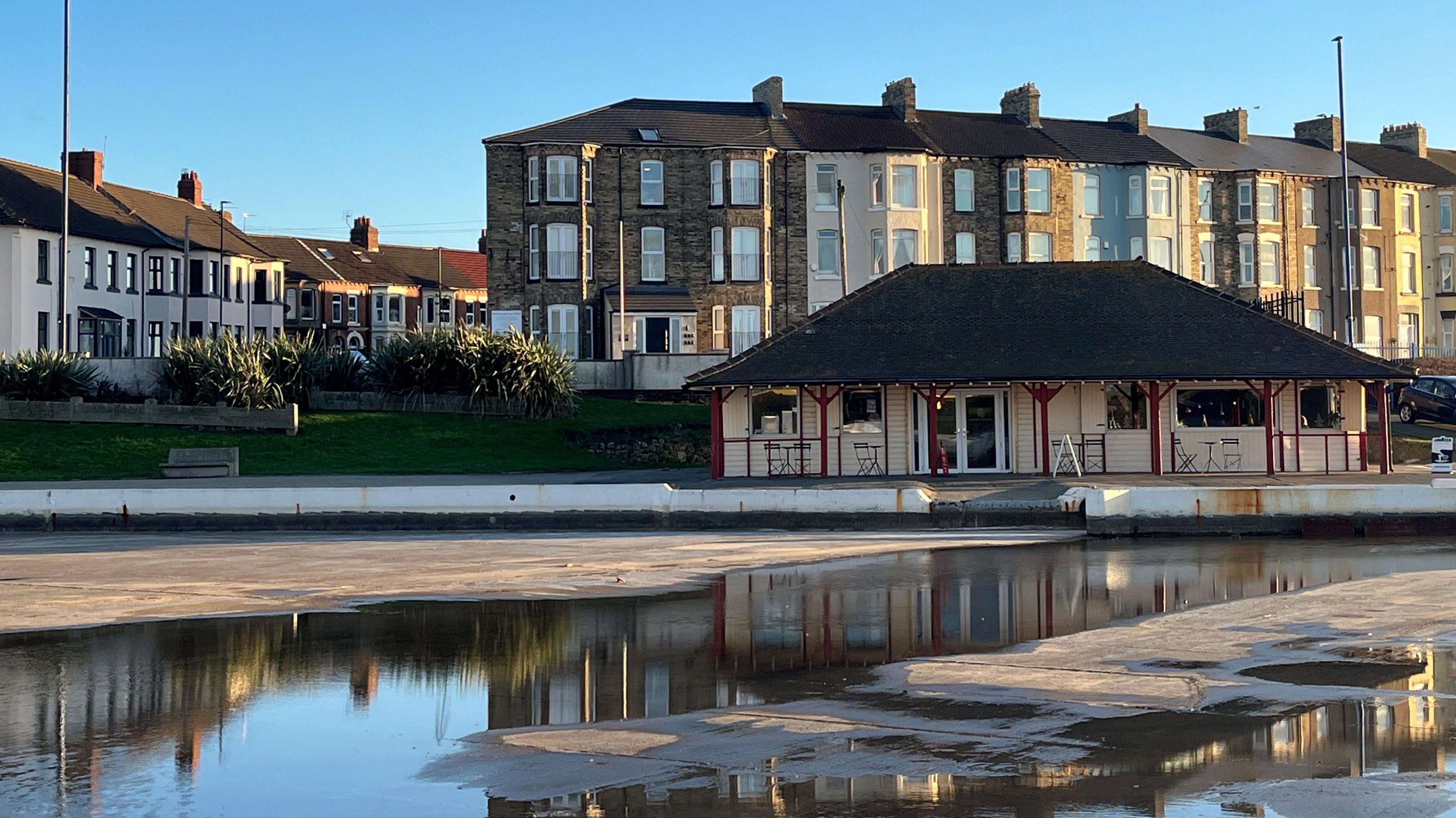 The Boathouse cafe. It is a small white house with black rood and red pillars.  Fairy lights are handing outside and there are tables and chairs.
The empty lake is in front of the building with some puddles from rain water in it. There are residential houses across the street behind the cafe. 