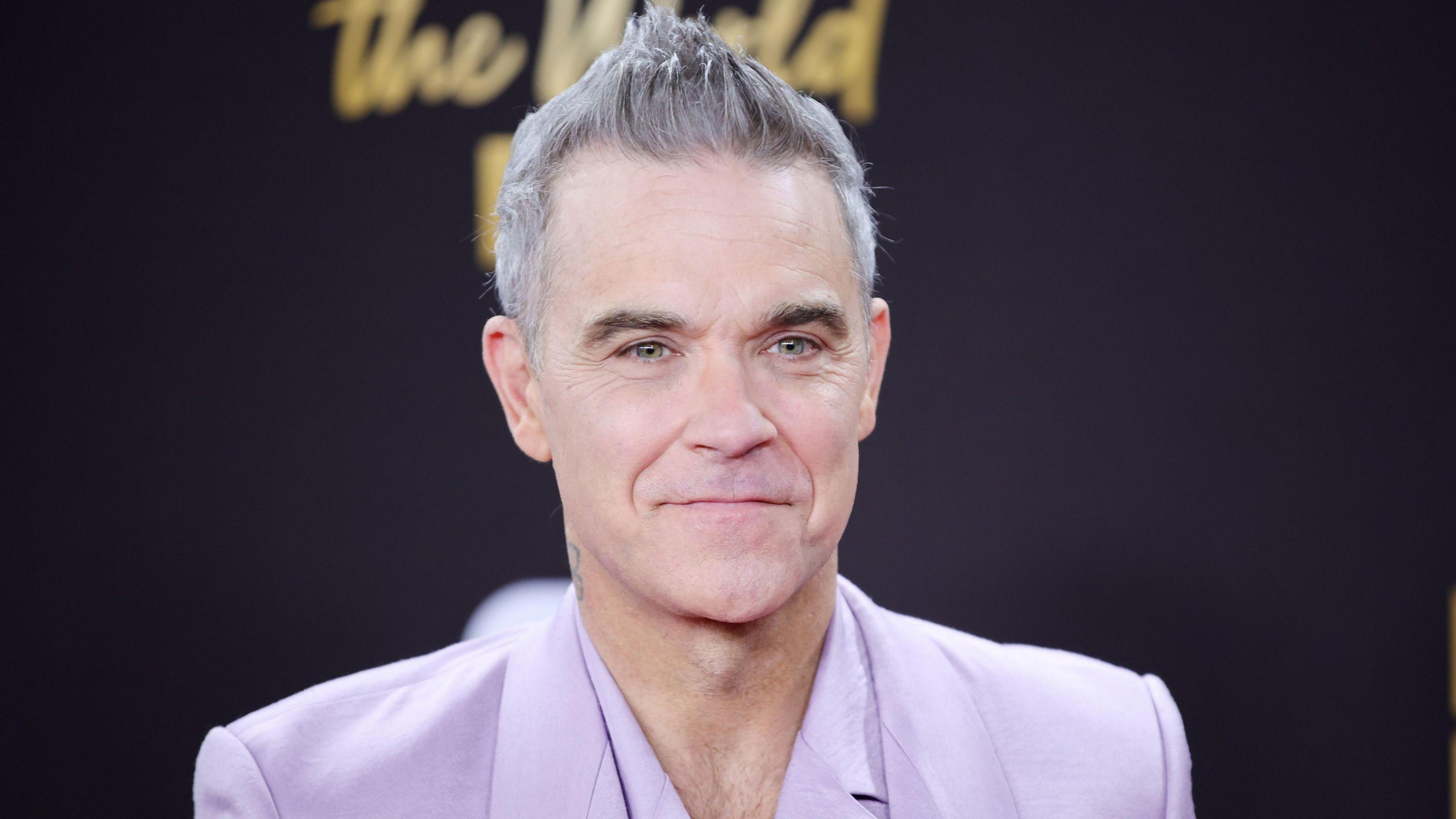 Robbie Williams, wearing a lilac-coloured suit, smiles as he stands in front of a black background.