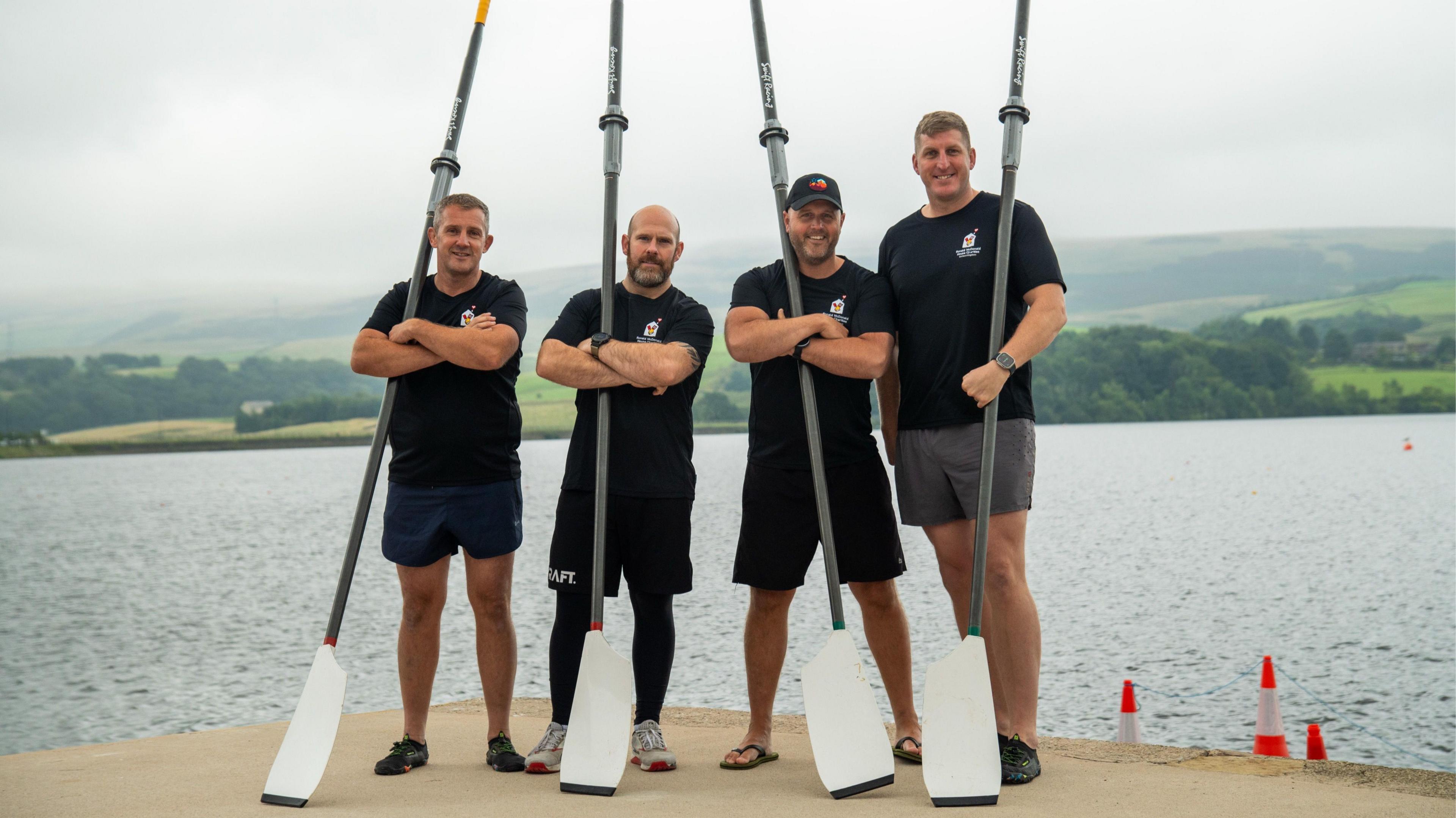 Four men in T-shirts and shorts stand on the edge of a lake carrying large oars.