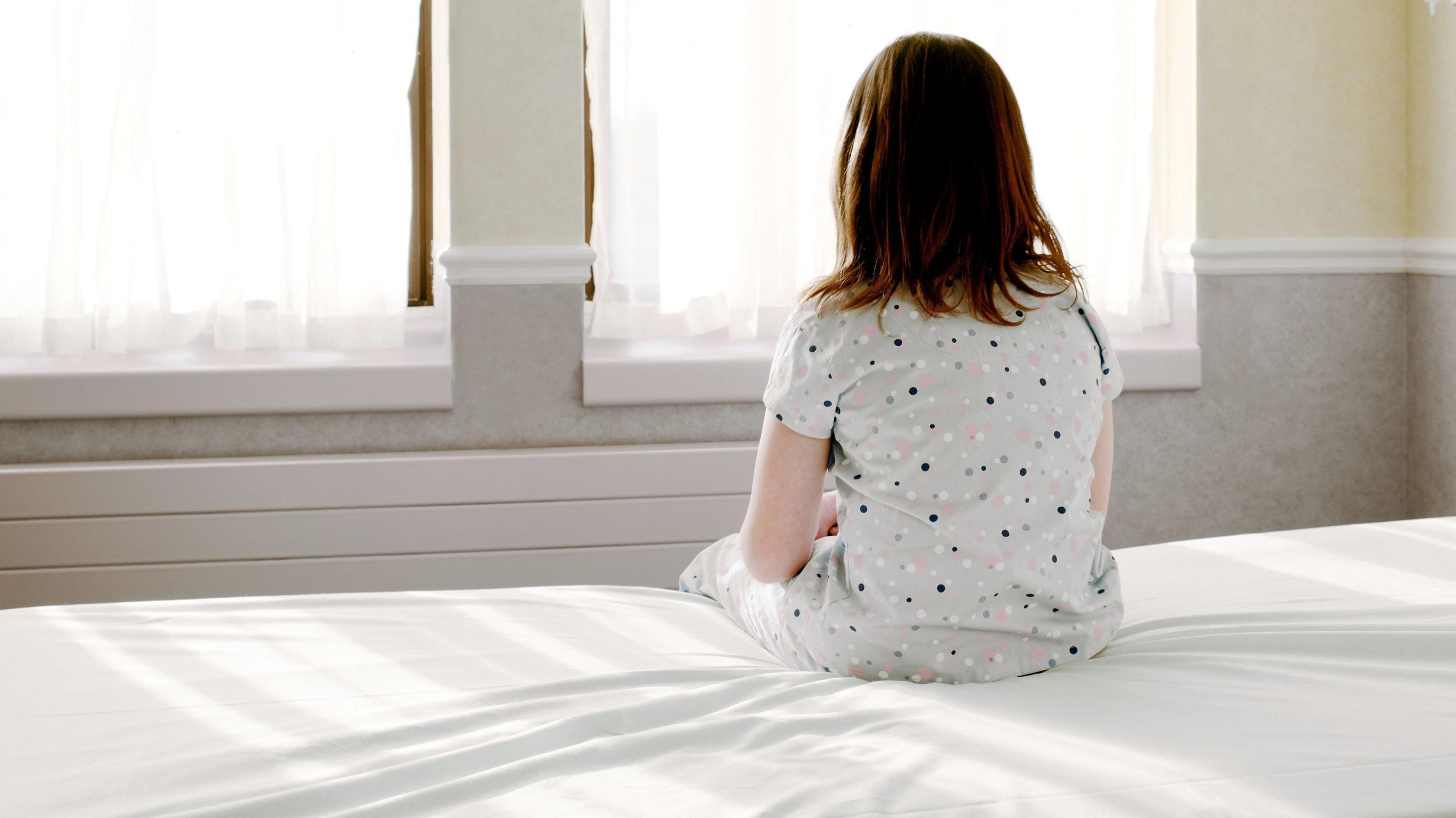 Young girl in nightdress sits on a bed with a white sheet with her back to the camera and looks out the window.