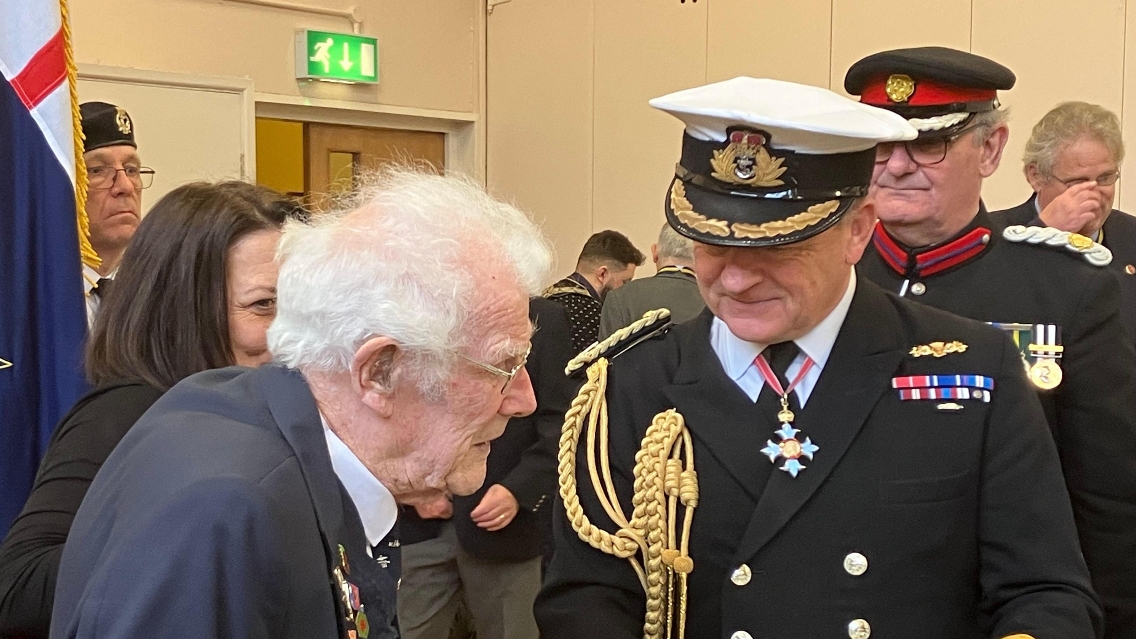 Jim Grant is wearing a suit and glasses and talking to Robert Bellfield who is wearing a formal military jacket with golden braid attached and a Royal Navy officer cap