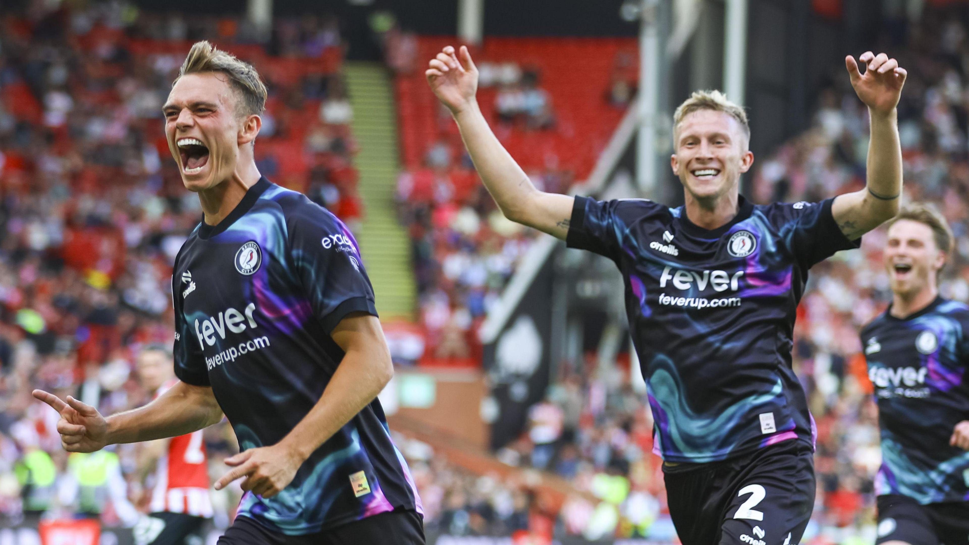 Scott Twine (left) runs with his mouth open smiling while pointing his finger with Ross McCrorie following behind with two arms in the air