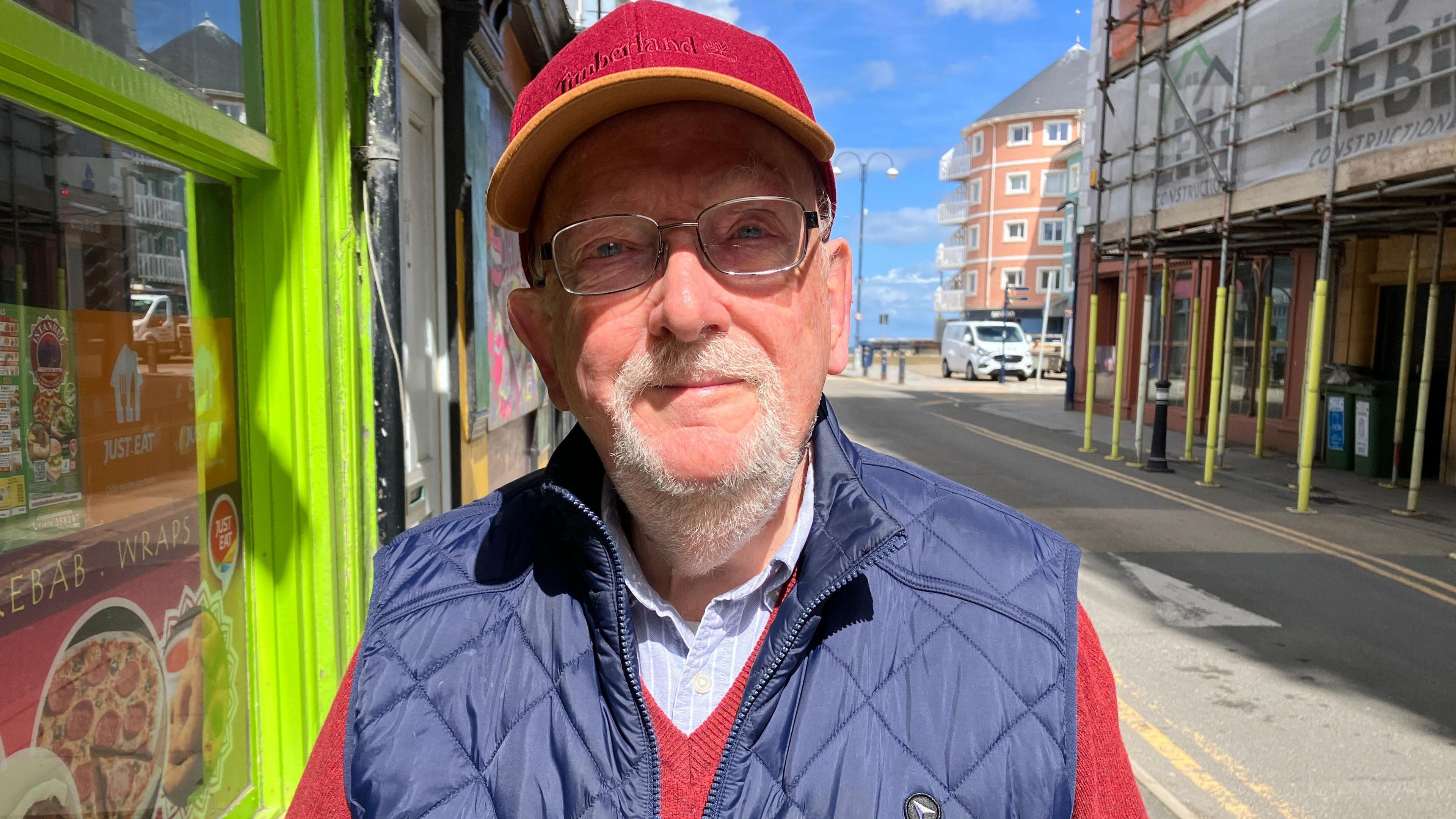 LLun o Geraint Evans yn nhref Aberystwyth - ei gefn at y prom, cap coch ar ei ben, sbectol a gilet glas