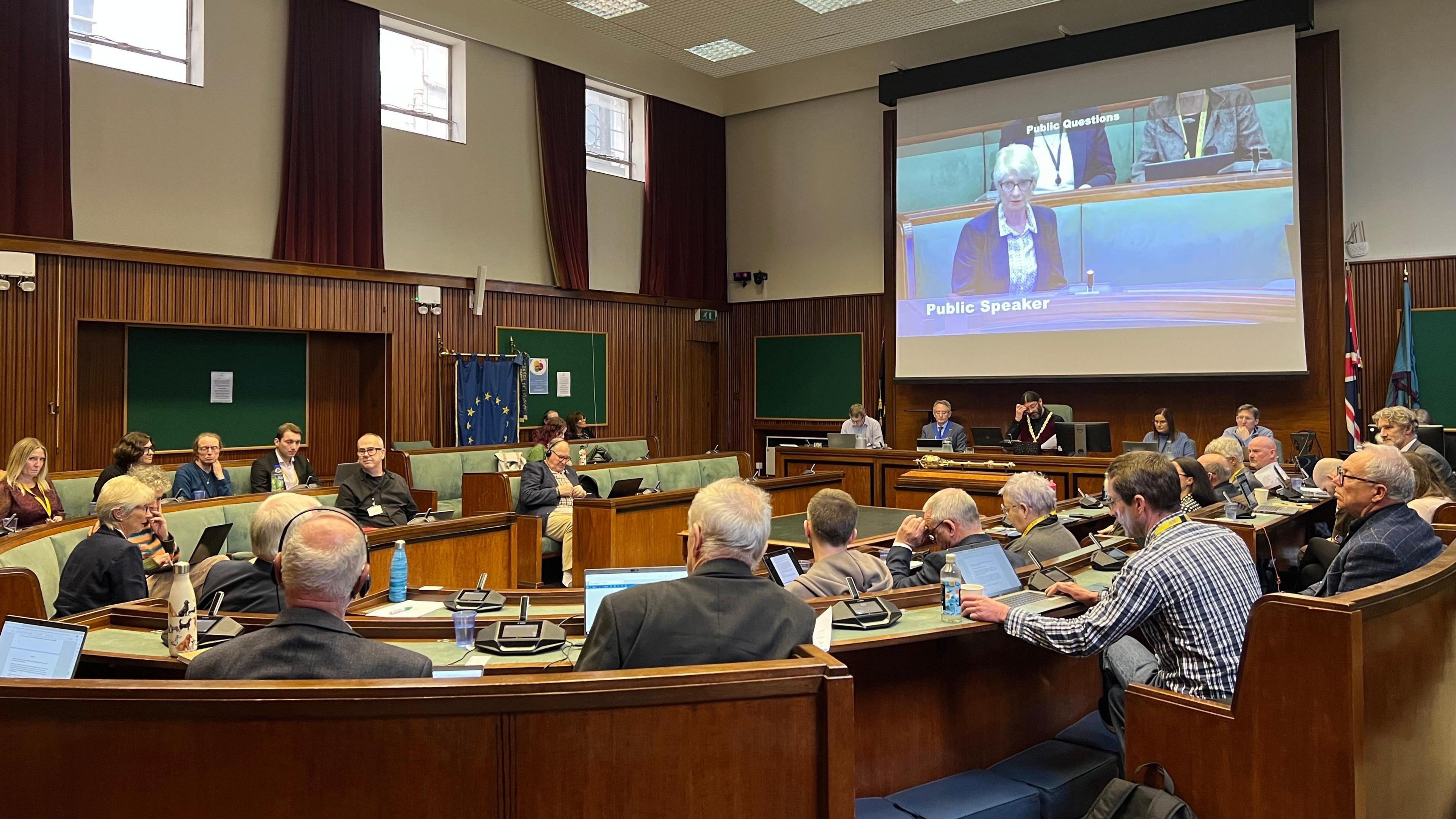 A woman with grey hair can be seen talking on a projector screen at a council meeting. Various councillors can be seen looking away from the camera with flags and windows visible in the background.