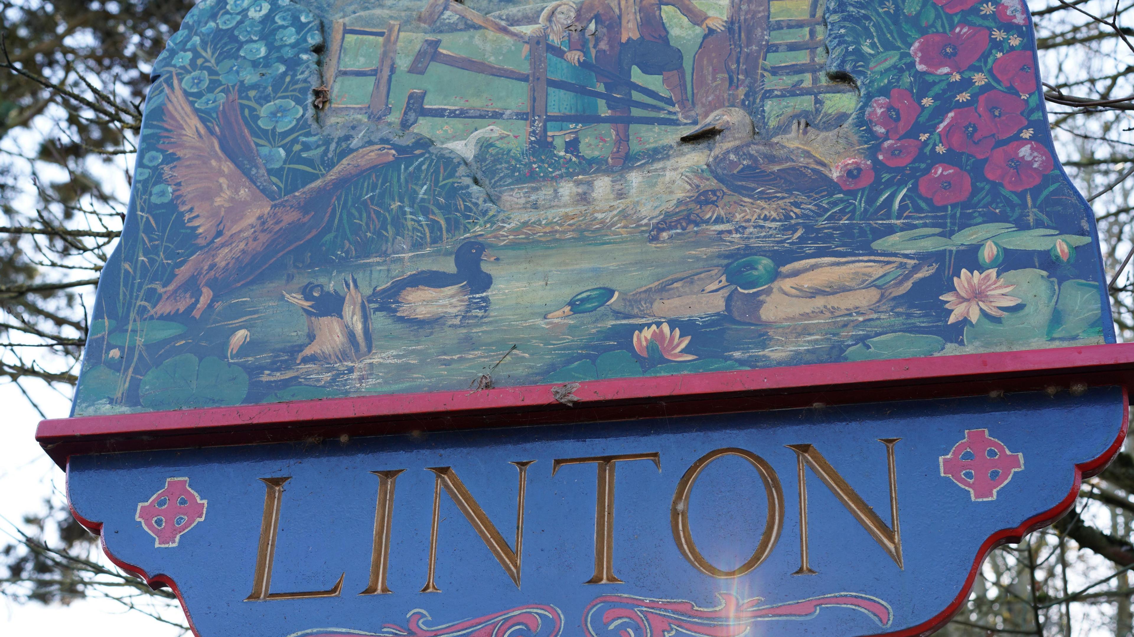 Sign for Linton, the village name is written in gold lettering, and the sign is painted with images of a river and birds.