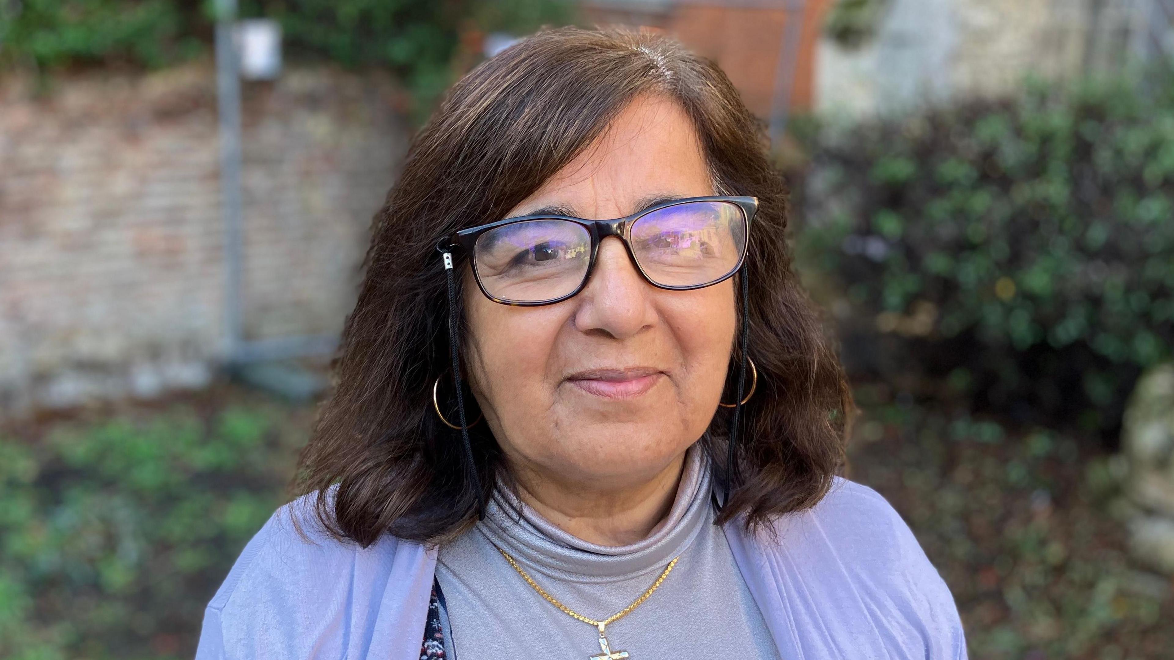 Asha is in the middle of the image looking at the camera. She had shoulder length brown hair and was wearing black framed glassed, a purple top and cardigan. 