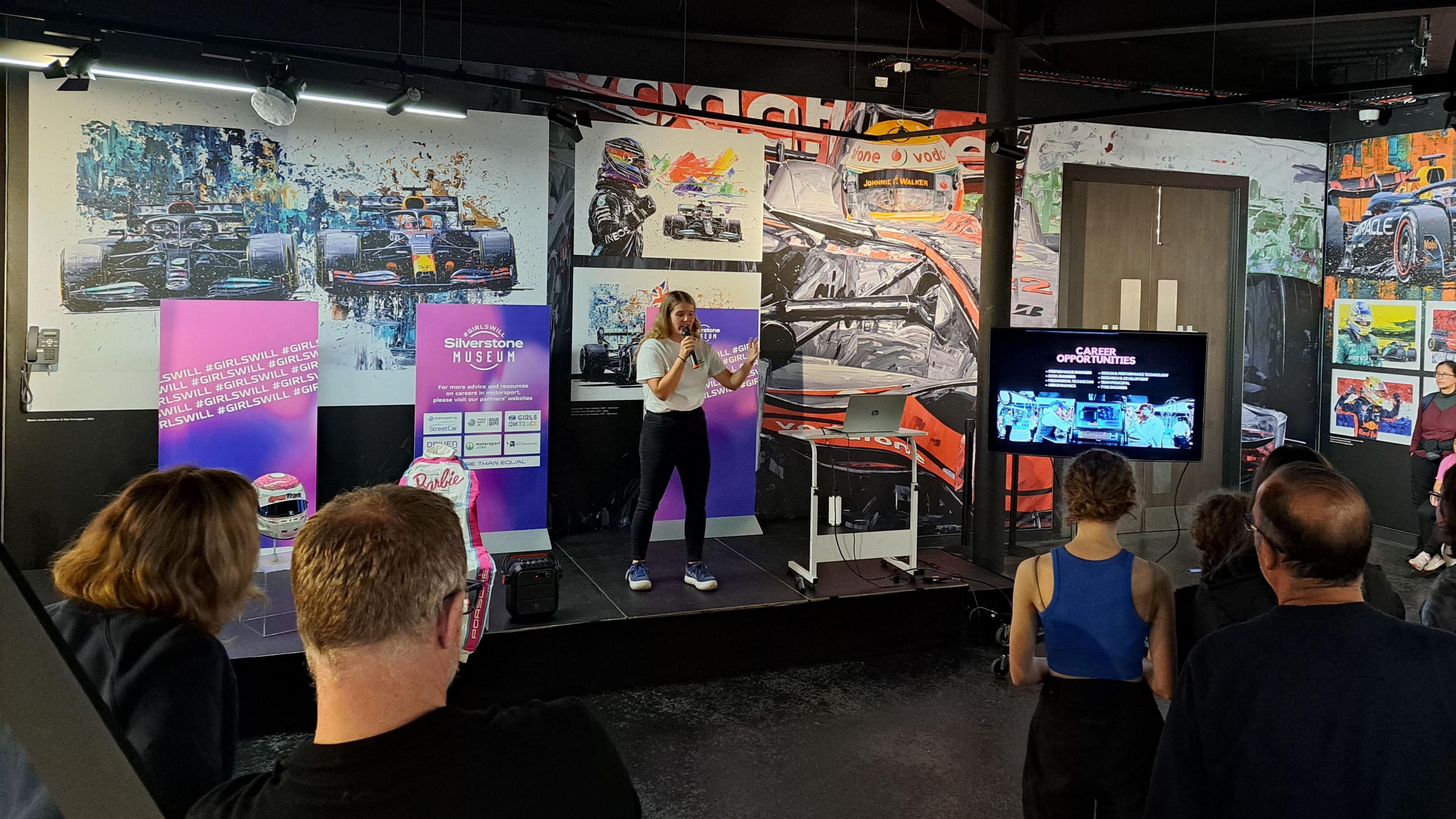 Racing driver Caitlin Wood is standing in front of racing car artwork and holding a microphone. In front of her is an audience sitting on chairs listening to her.