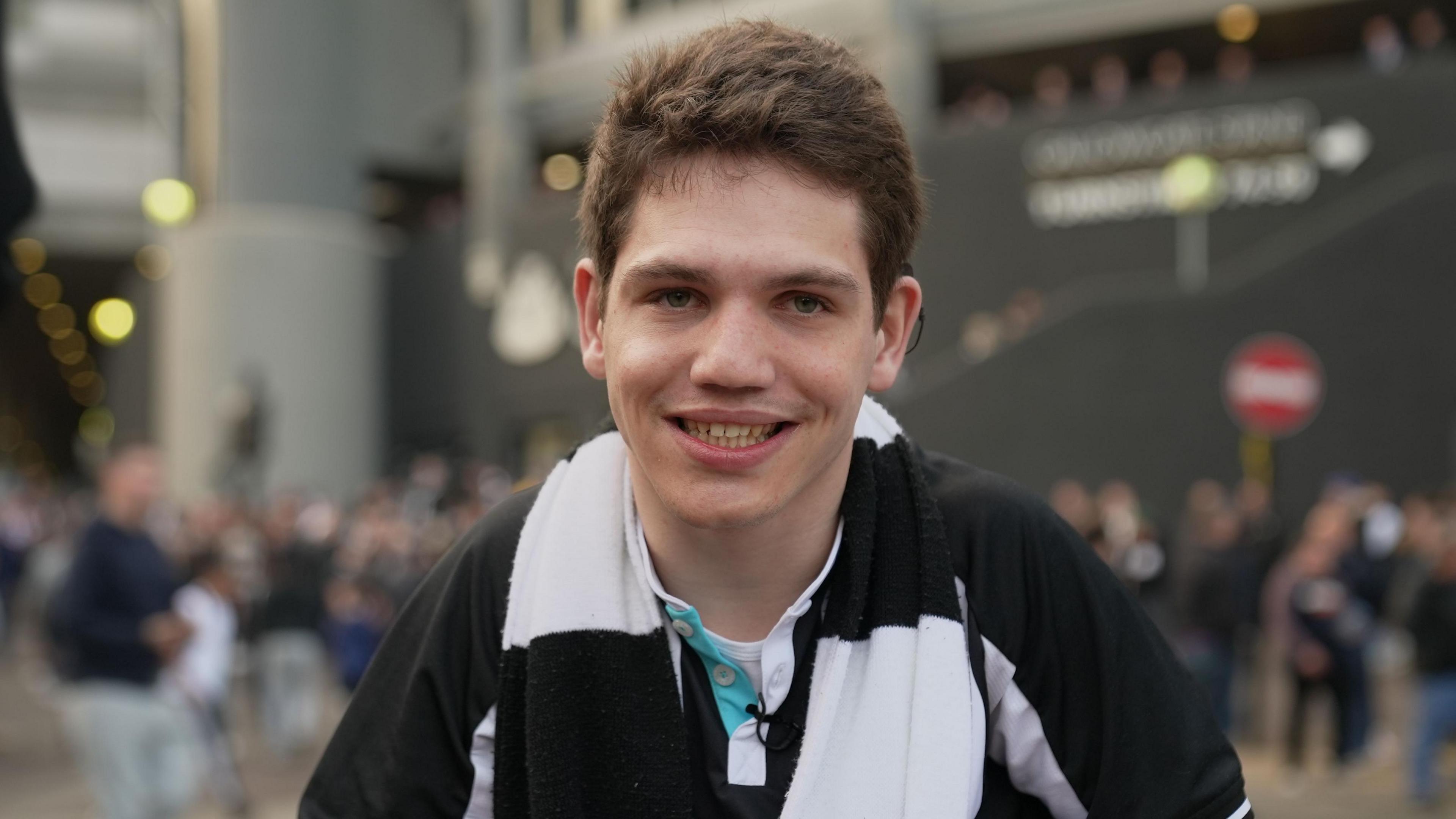 Elliot in his Newcastle United kit, wearing a black and white scarf. He is in front of St James' Park stadium and smiling at the camera.