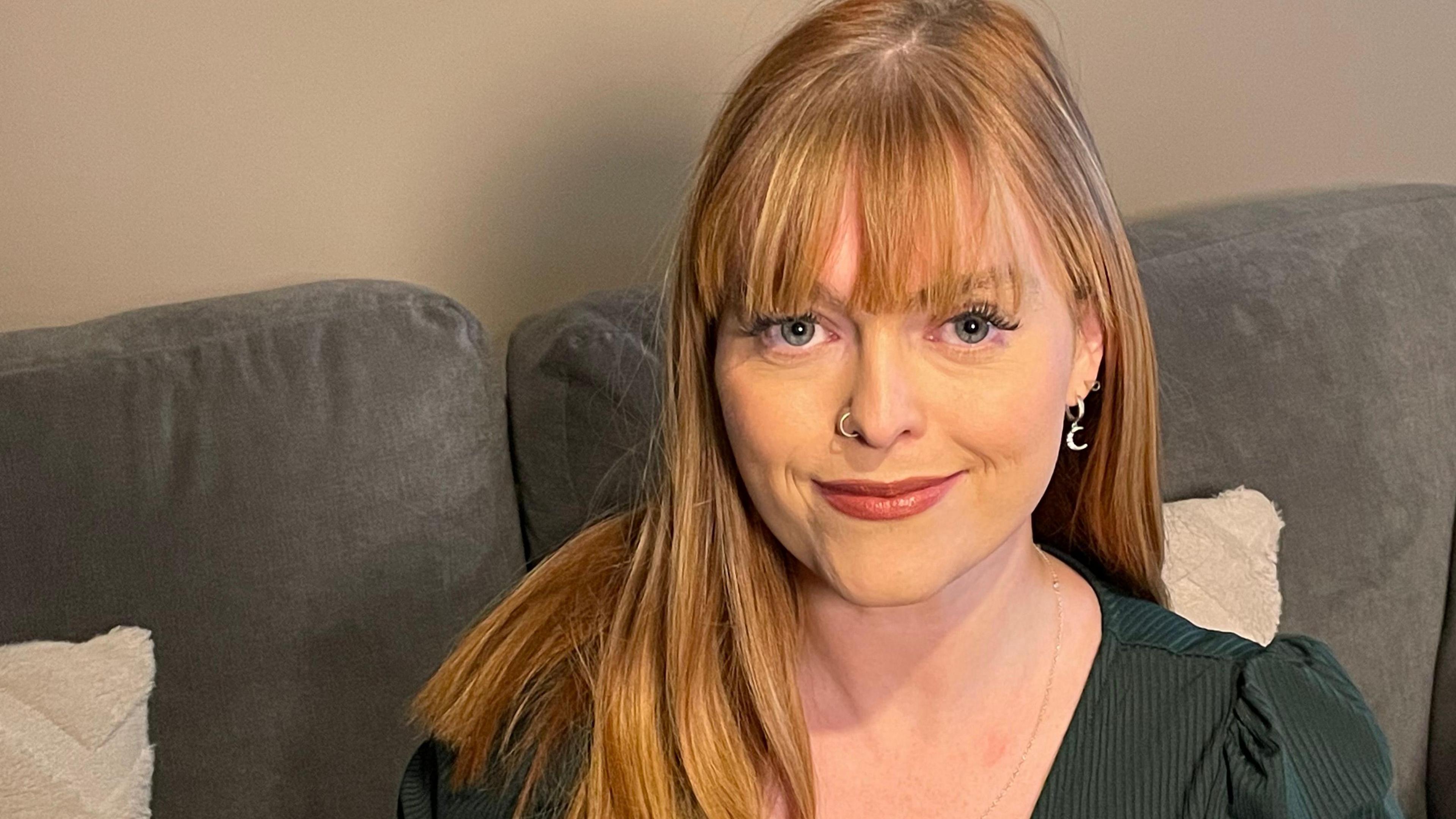 Sarah Curtiss, a woman with long ginger hair, smiles as she sits on a grey sofa. She is wearing a green top and has a silver nose ring and silver earrings.