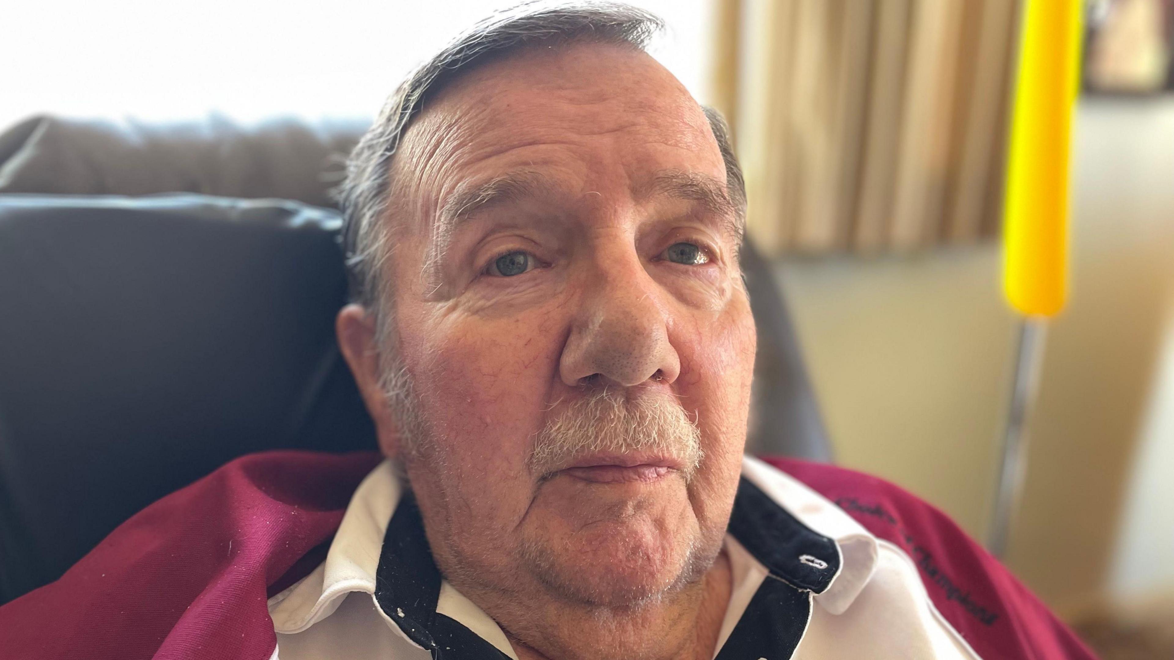 Russell sat in an armchair. He has short grey hair and has a moustache.