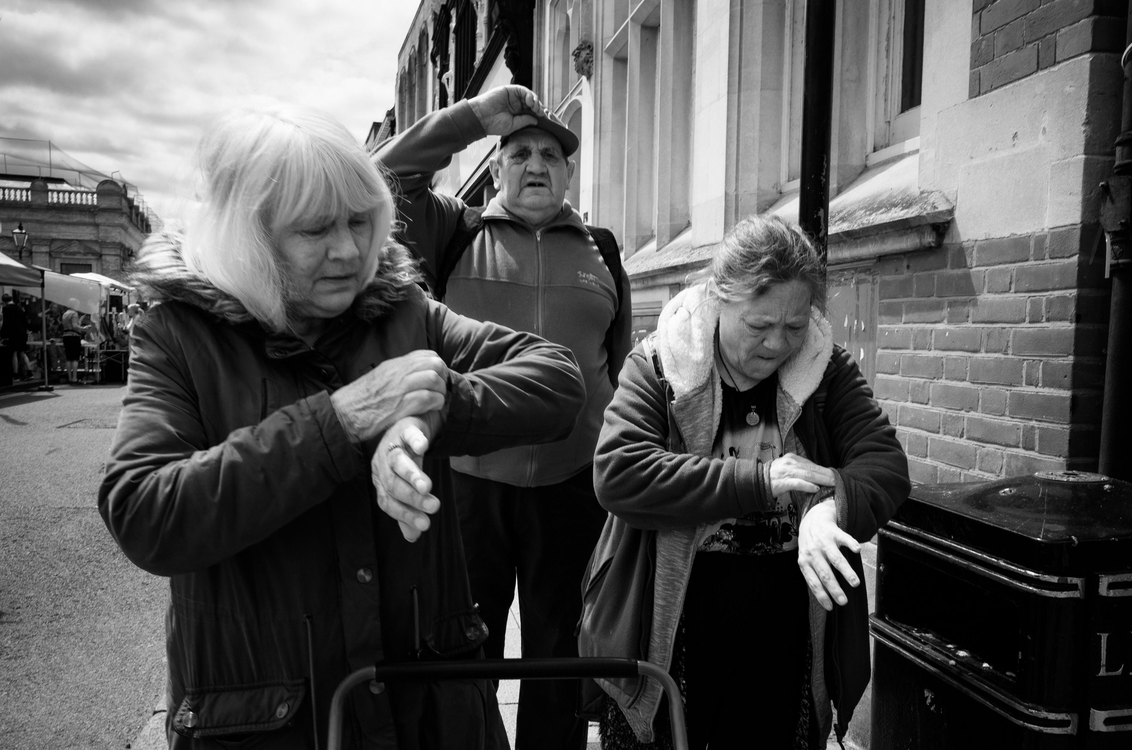 Two women check their watches, both wearing them on their left wrists. A man stands behind them looking towards the camera, with one hand on his flatcap, appearing to raise it slightly from his head