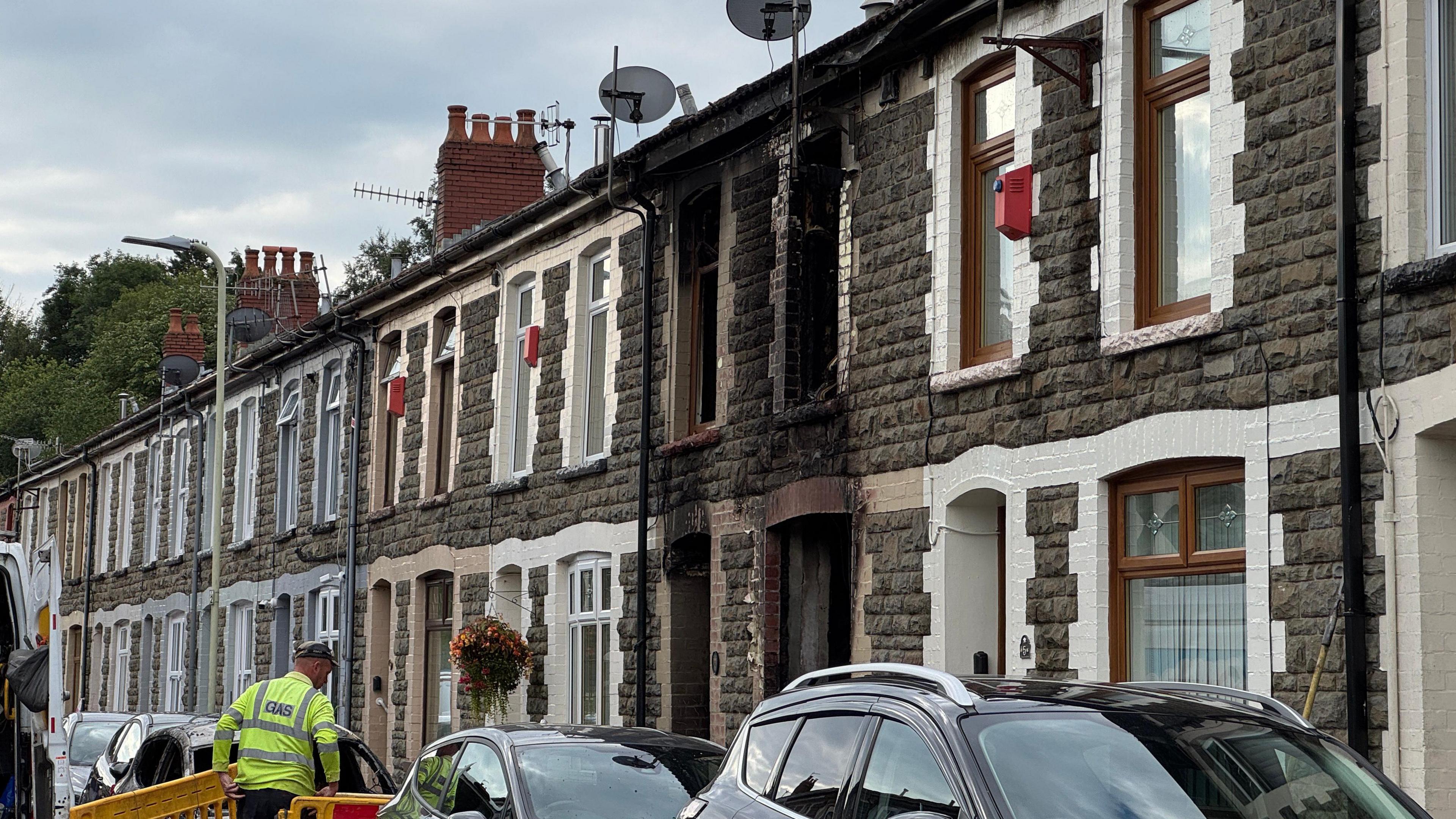 A house with fire and smoke damage. It sits in the middle of a row of terraced houses on a street. There are cars parked outside of the houses. A man wearing a neon yellow jacket is placing barriers around the house.